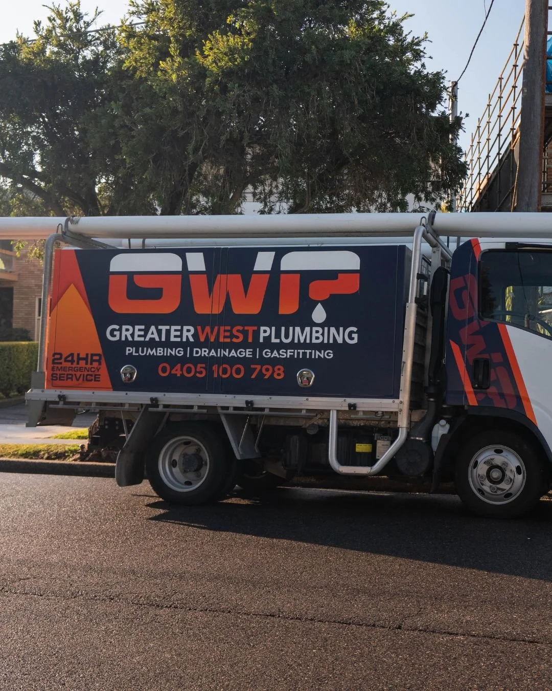 A Greyhound plumbing truck parked on a street, with trees and a building in the background, displaying advertisement for Greater West Plumbing services.