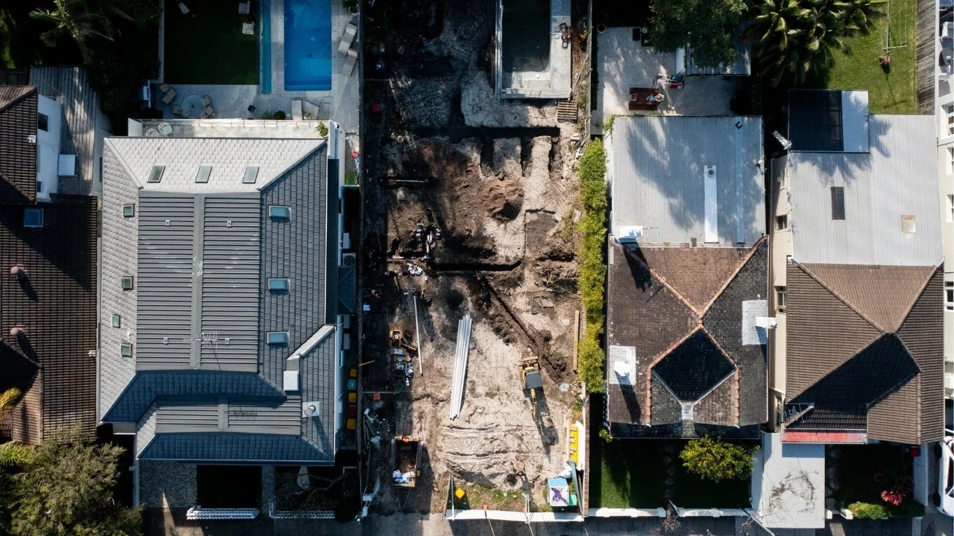 Aerial view of a construction site between two residential houses, with excavated dirt and construction equipment.