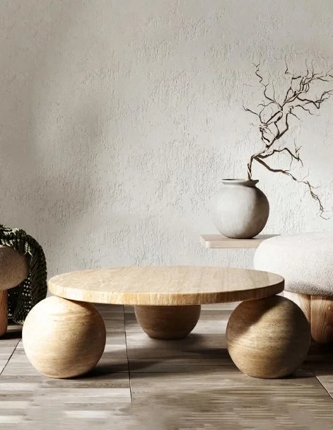 Modern living room with a round wooden coffee table on three spherical legs, a beige sofa with a white cushion, a white wall with a decorative shelf holding a ceramic vase with dried branches, and hardwood flooring.