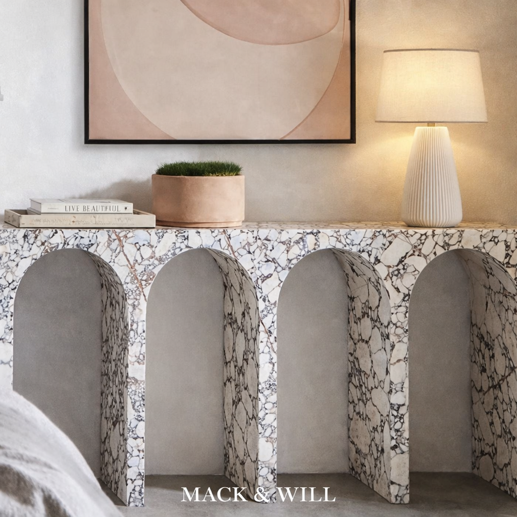 A modern marble console table with a black and white pattern, against a neutral wall. On top of the table, there are two stacked books with the titles "Live Beautiful" and another book, a round pink planter with green grass, and a cream-colored table lamp with a ribbed base and lampshade. Above the console, there is a large abstract artwork with soft, muted tones. The brand name "MACK & WILL" is displayed at the bottom of the image.