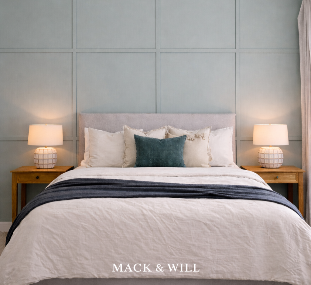 A neatly made bed with a light gray upholstered headboard, white bedding, a dark blue throw blanket, and multiple pillows including one dark blue pillow, flanked by wooden nightstands with white table lamps on each side. The wall behind the bed has a gray paneled design. The words "MACK & WILL" are written at the bottom of the image.