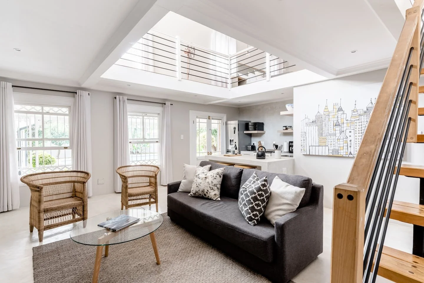 Living room with a gray sofa, wicker chairs, a glass coffee table, and a kitchen area in the background, featuring a white counter, black appliances, and a cityscape artwork on the wall. Sunlight enters through multiple windows with white curtains.