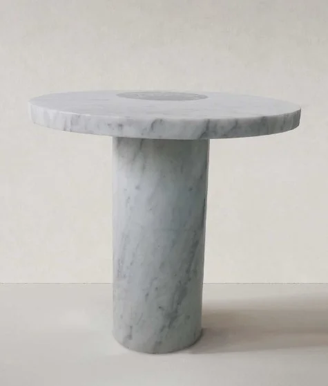 A round, white marble table with a single cylindrical marble pedestal base, set against a plain light-colored background.