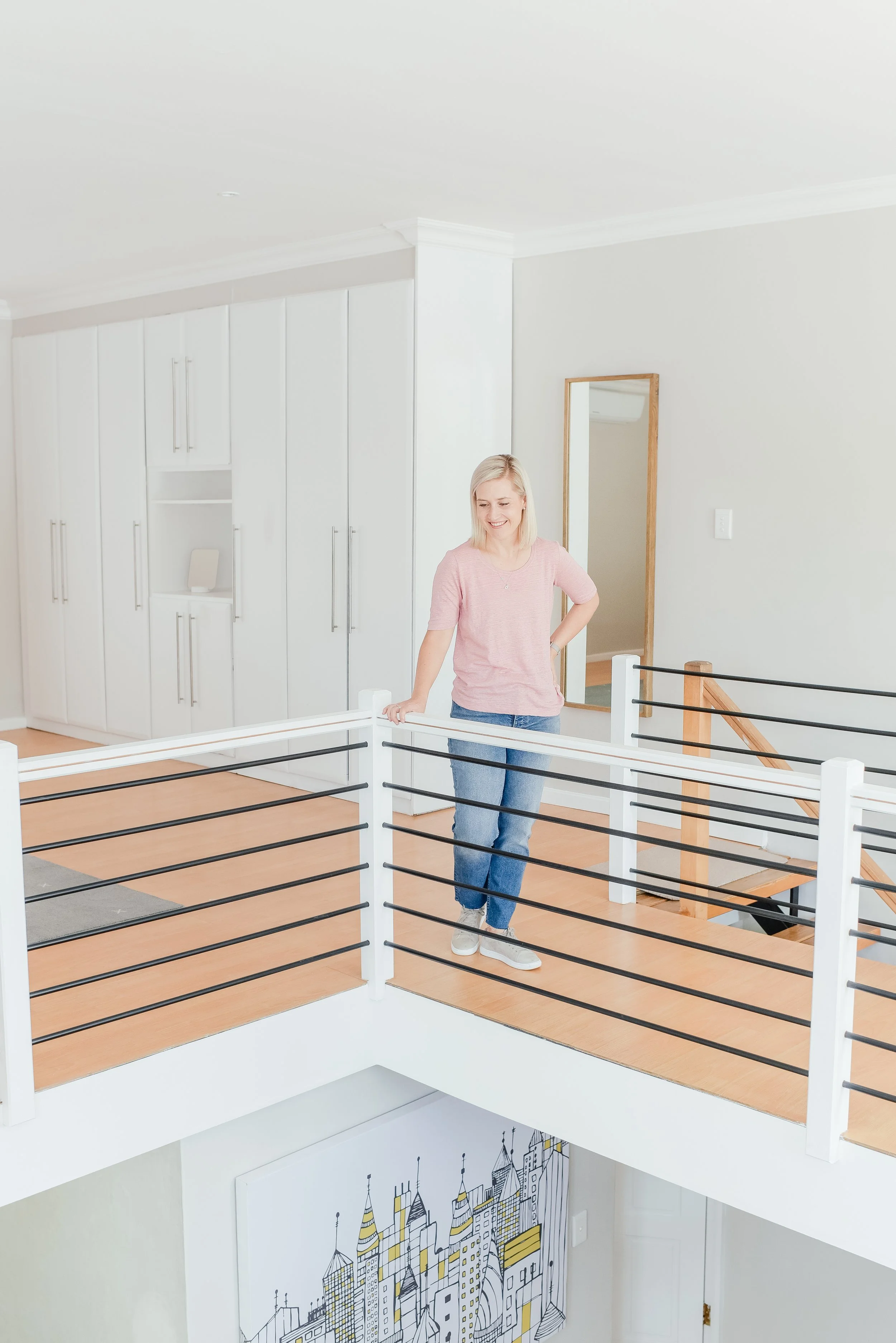 A woman in a pink shirt and jeans smiling and leaning on a white railing on an upstairs landing in a modern, bright home with white walls and built-in cabinets.
