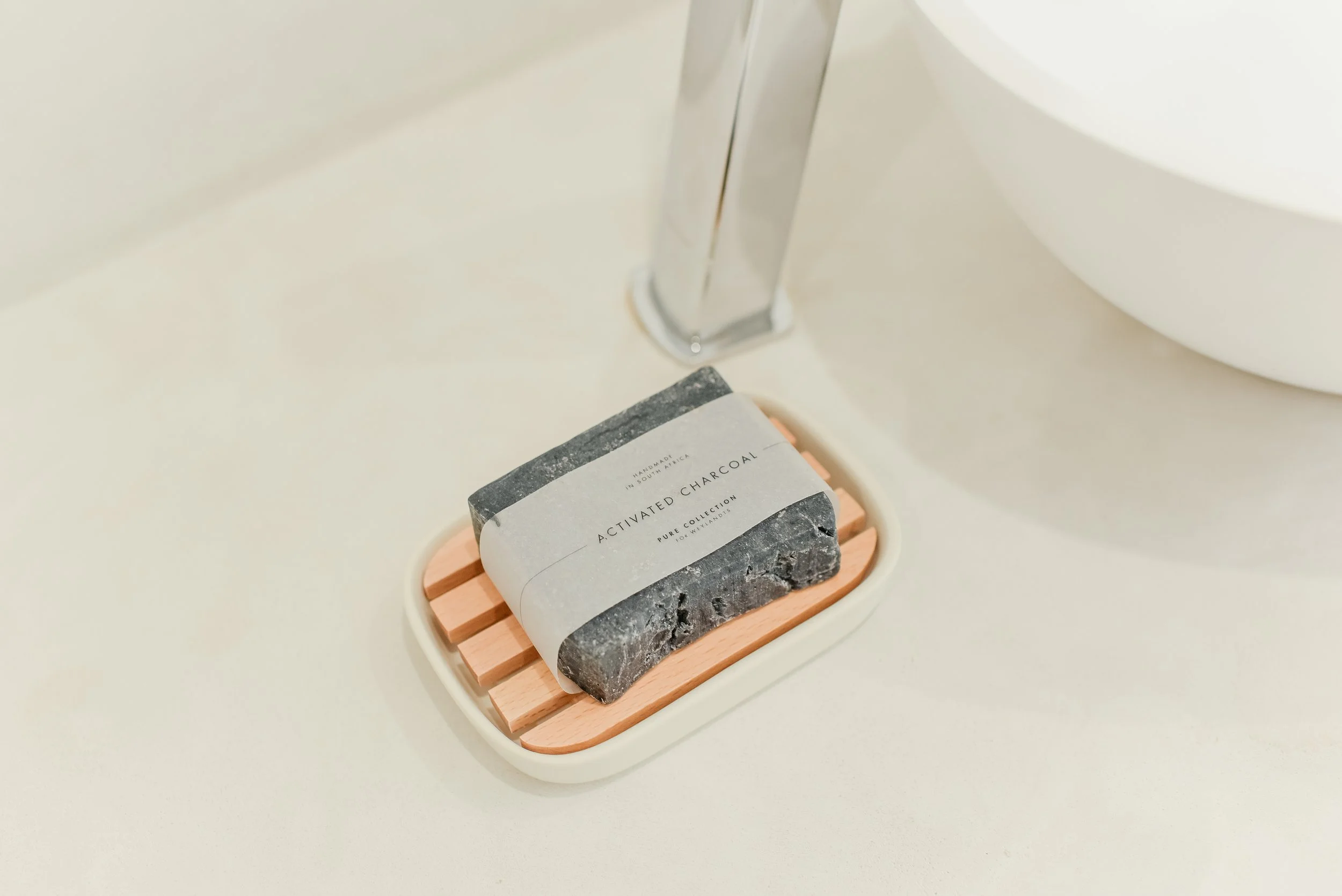 A bar of activated charcoal soap with a textured dark gray surface and a paper band, placed on a wooden soap dish on a marble countertop.
