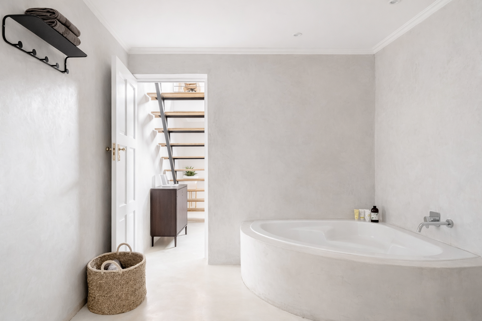 A minimalist bathroom with a corner bathtub, a woven basket on the floor, a wall-mounted shelf with towels, and a doorway leading to a room with a staircase and side table with plants.