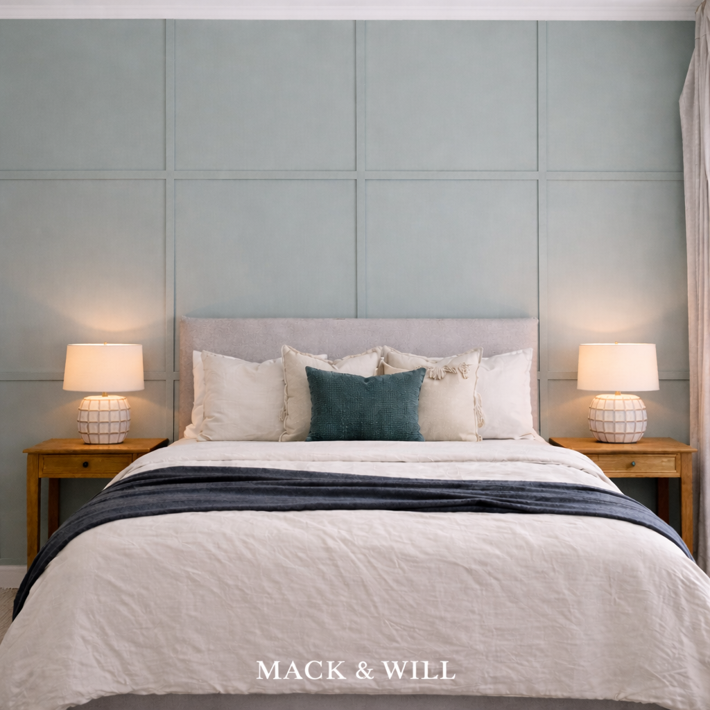 A neatly made bed with a light grey padded headboard, white bedding, and multiple cream and dark teal pillows, flanked by two wooden nightstands with matching lamps, against a light blue paneled wall.