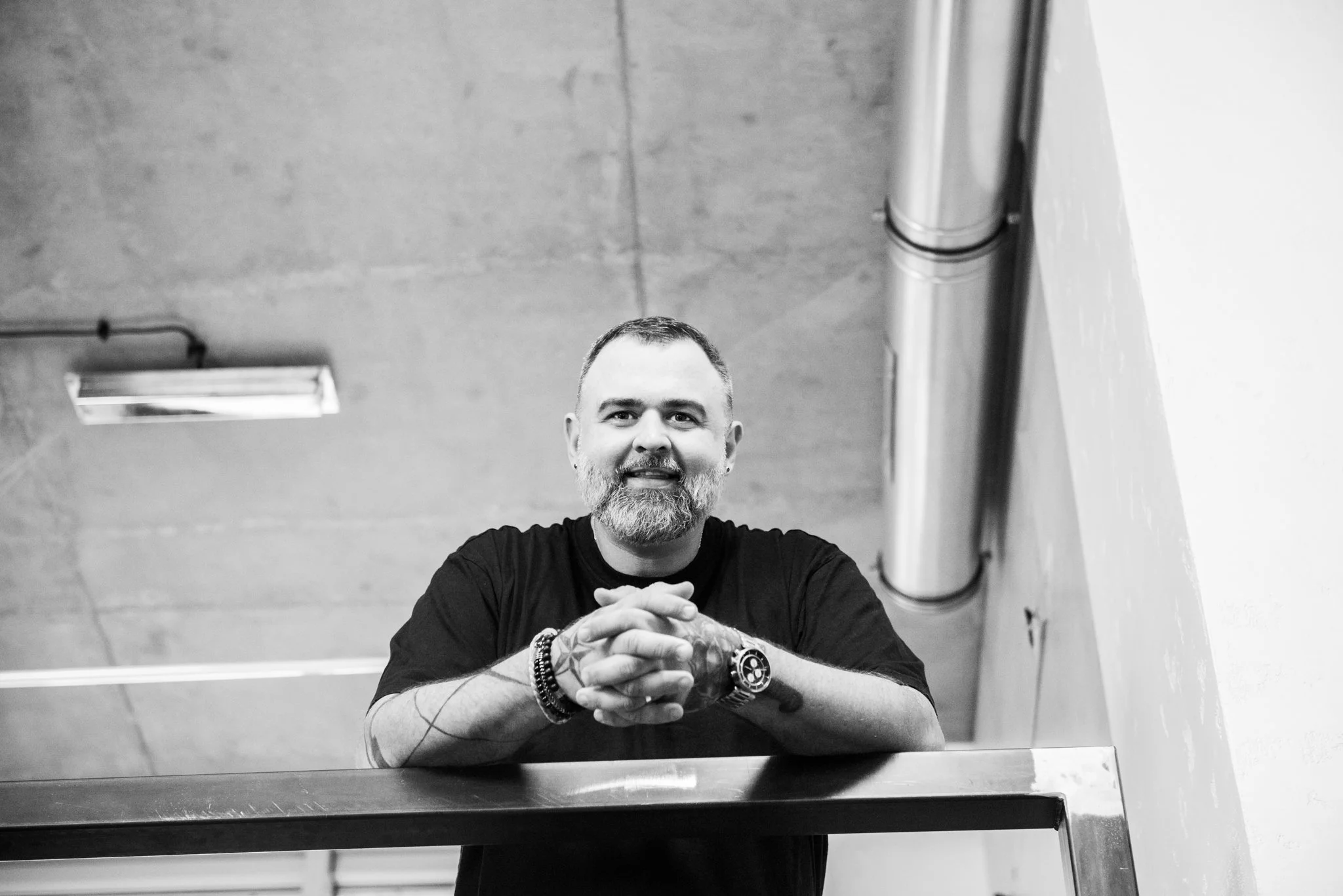 A man with a beard and short hair, wearing a black t-shirt and bracelets, sitting at a table and smiling at the camera in front of a mirror.