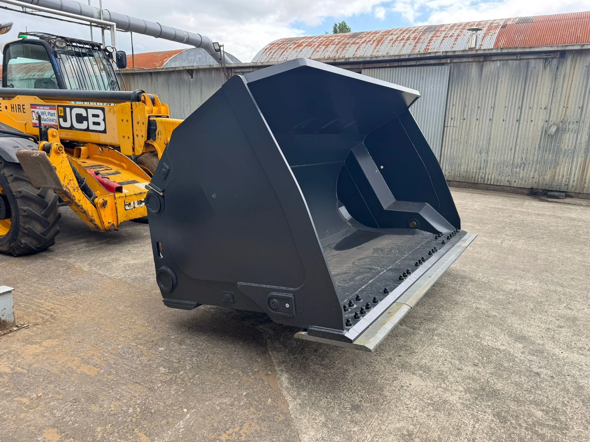 A large, black excavator bucket attachment rests on the ground next to a yellow JCB compact loader, with a corrugated metal building in the background.