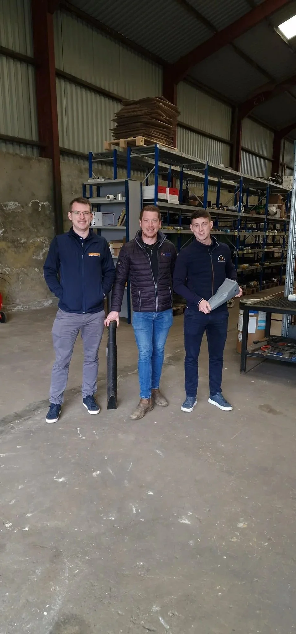 Three men standing inside a warehouse with metal shelves and concrete walls, one holding a large black hydraulic chisel attachment, another holding a gray bucket tooth, smiling at the camera.