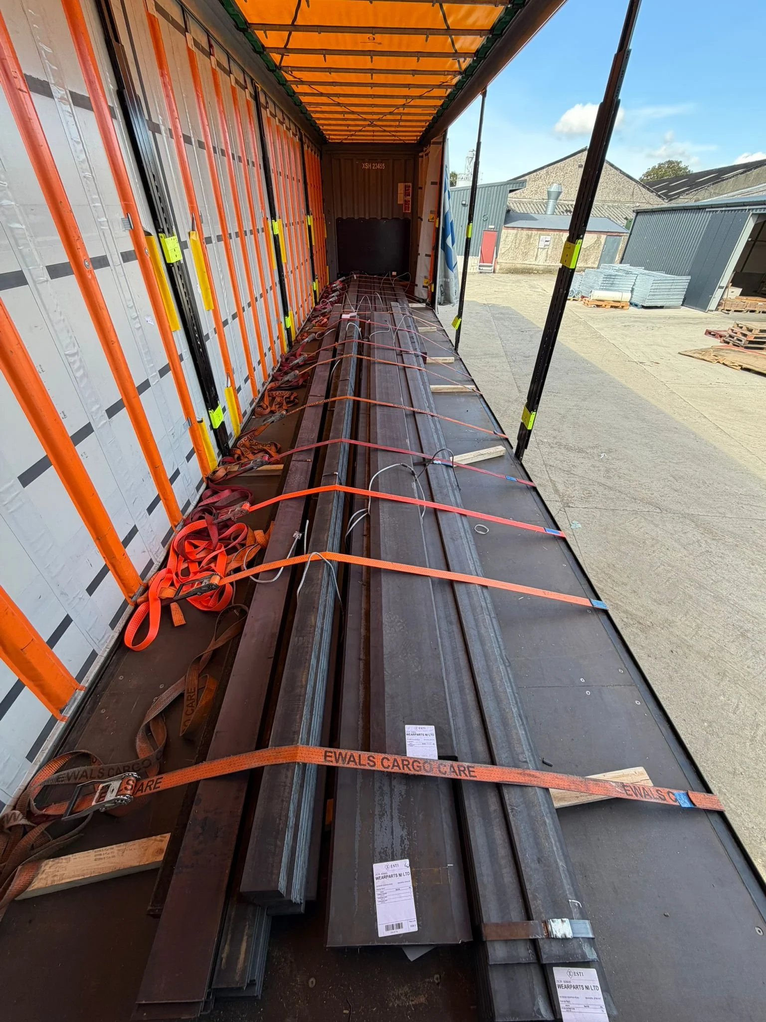 Steel bars and beams secured with orange straps inside a transportation container on a flatbed truck.