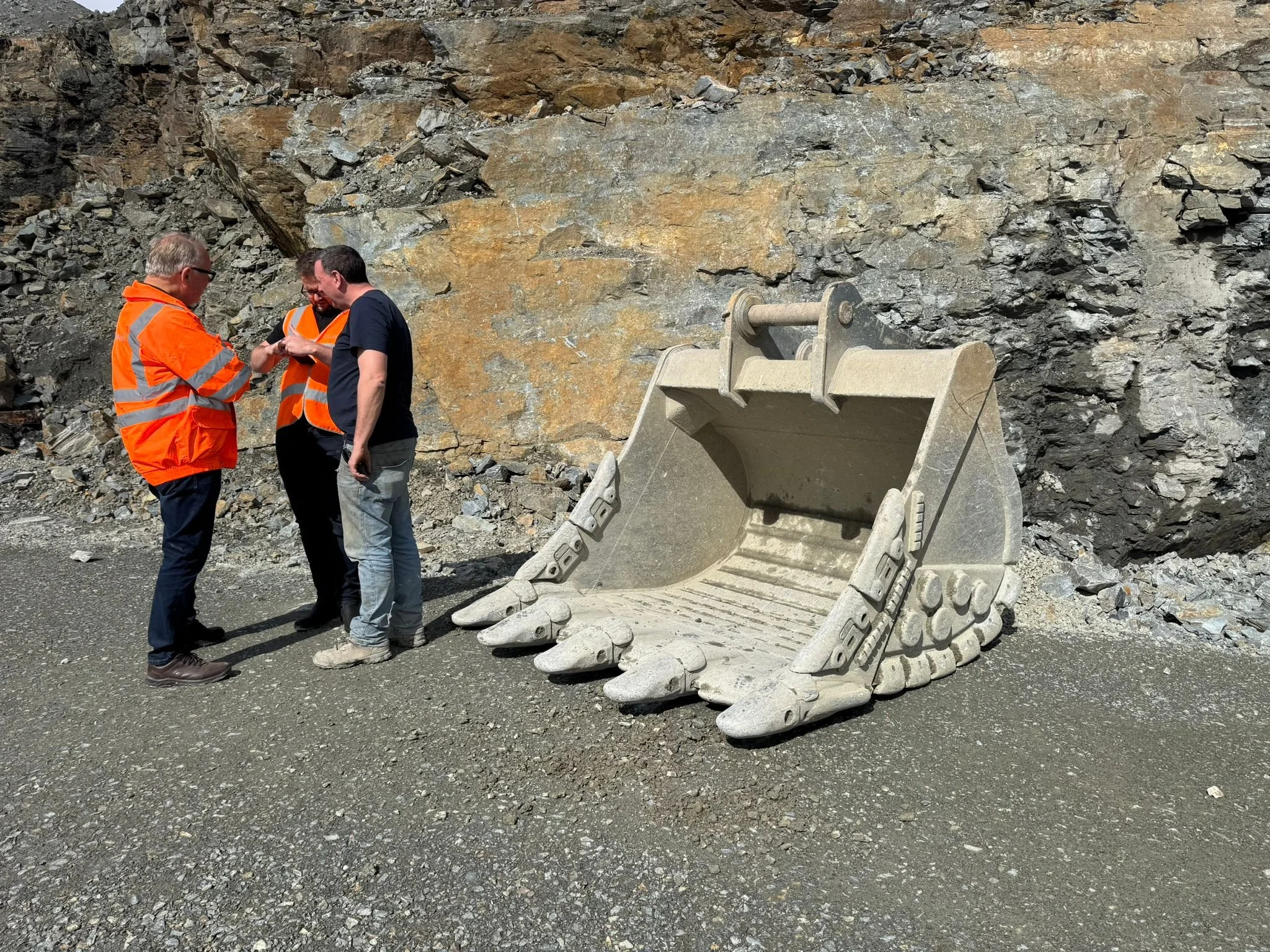 Three men are standing together on a gravel surface in front of a rocky cliff. Two of them are wearing orange safety vests. They are examining or discussing something. Next to them, an excavator bucket is resting on the ground, showing detailed teeth