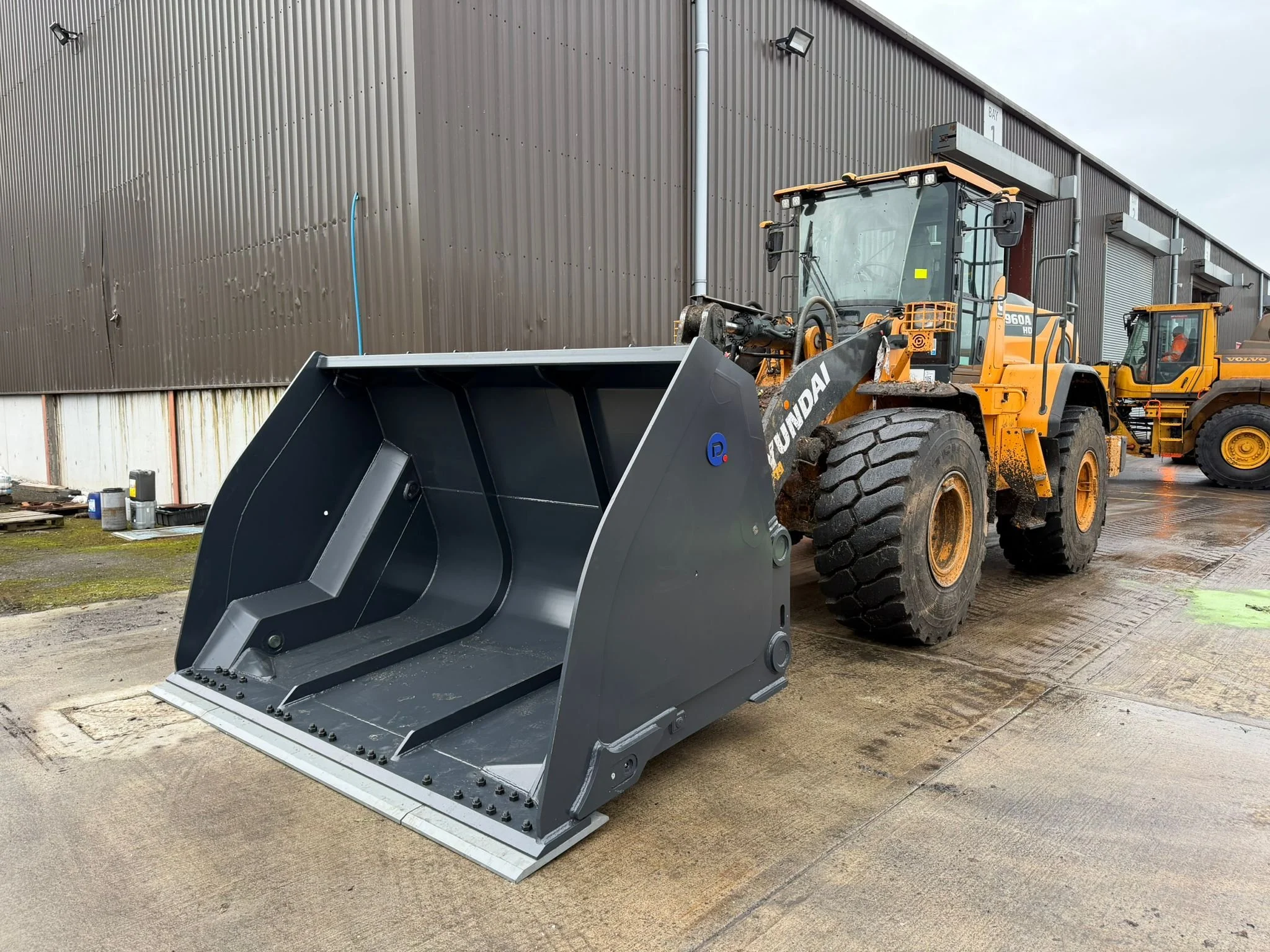 Yellow Hyundai wheel loader with black bucket attachment parked outside industrial building with dark metal siding. Another construction vehicle visible in the background. The ground is wet.
