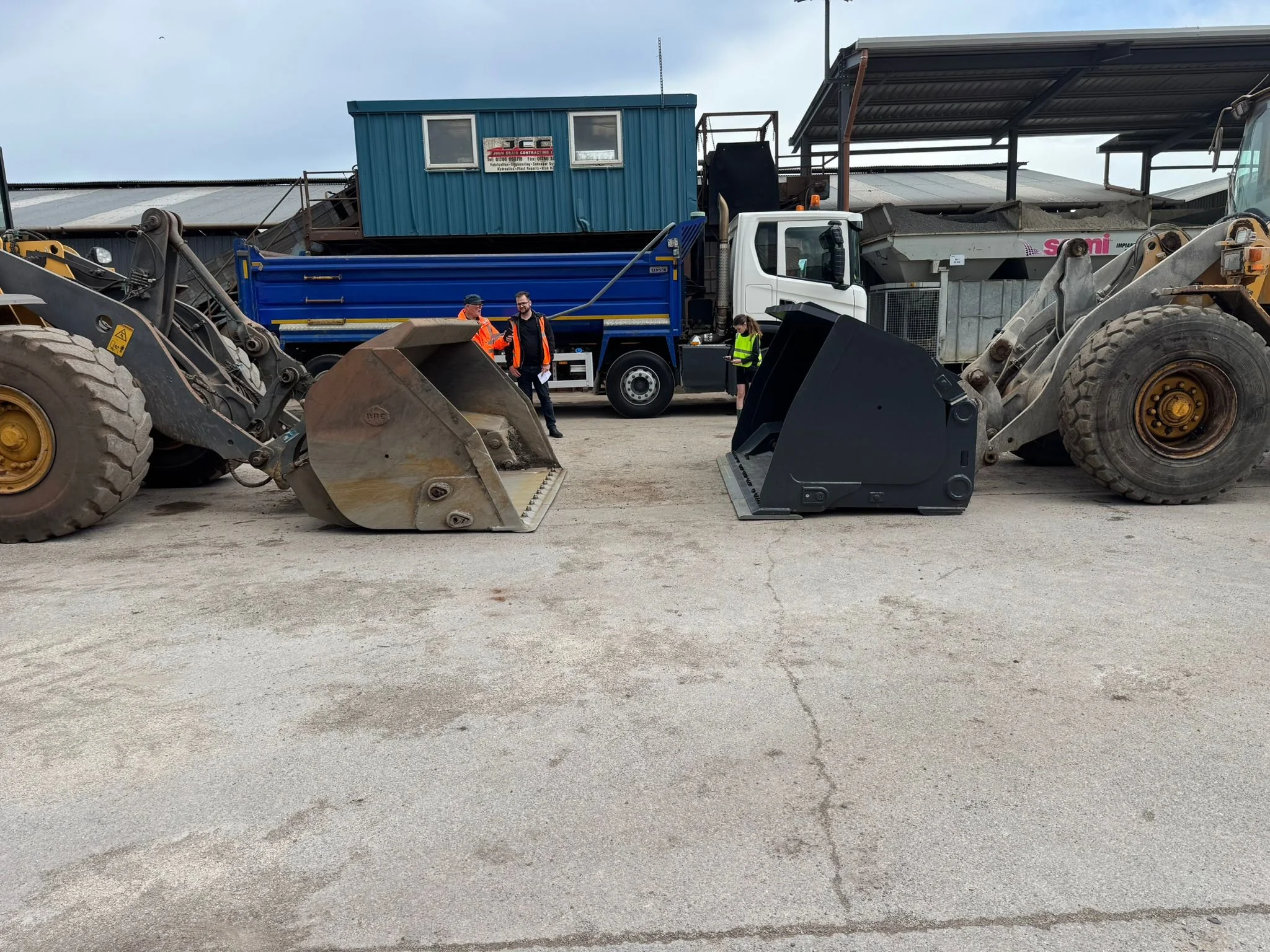 Construction yard with two large excavators facing each other, and a blue truck in the background. Several workers in safety vests are talking near the truck.
