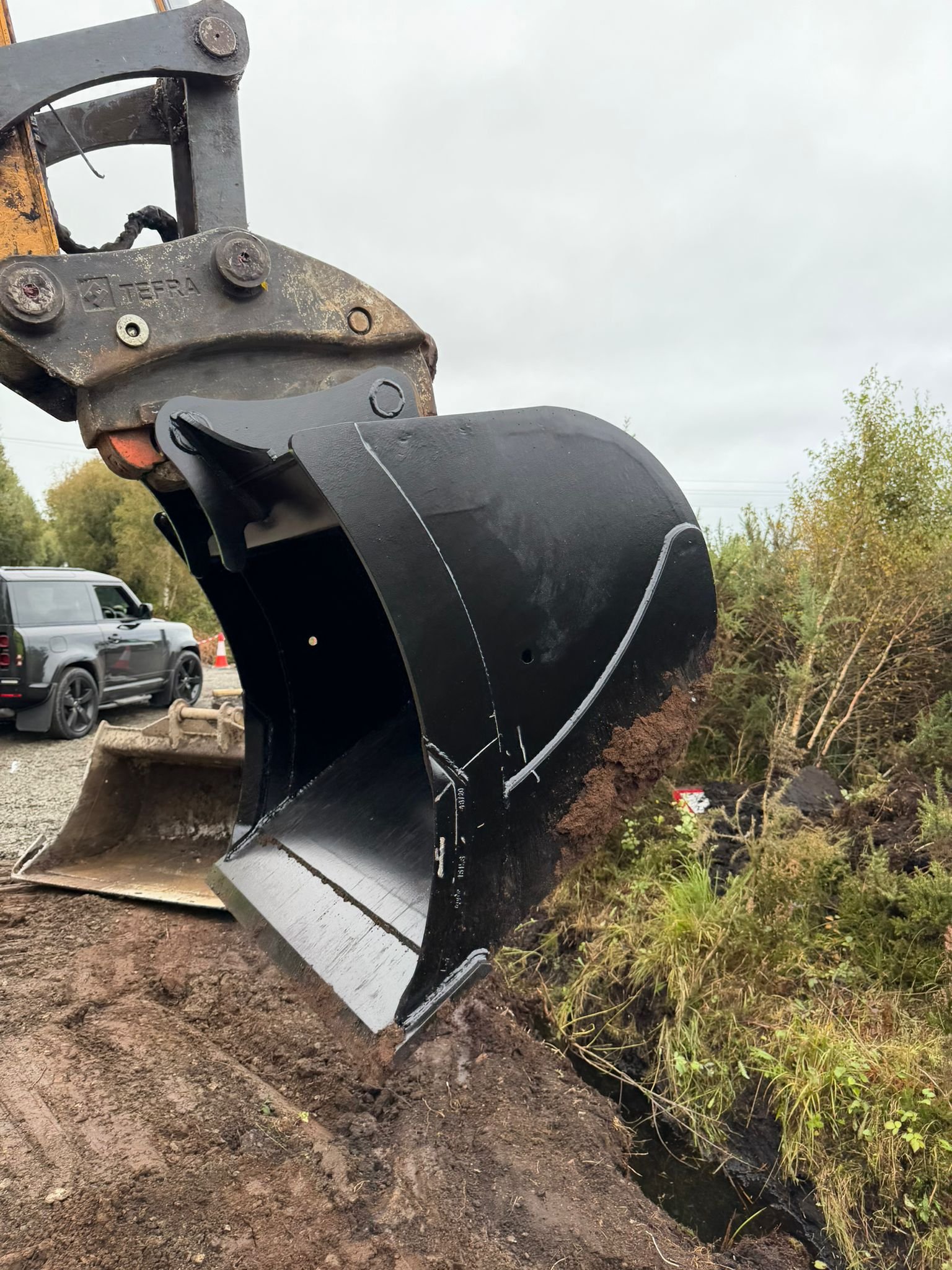 Close-up view of a large black excavator bucket attachment on construction equipment, digging into the soil on a cloudy day with trees and a vehicle in the background.