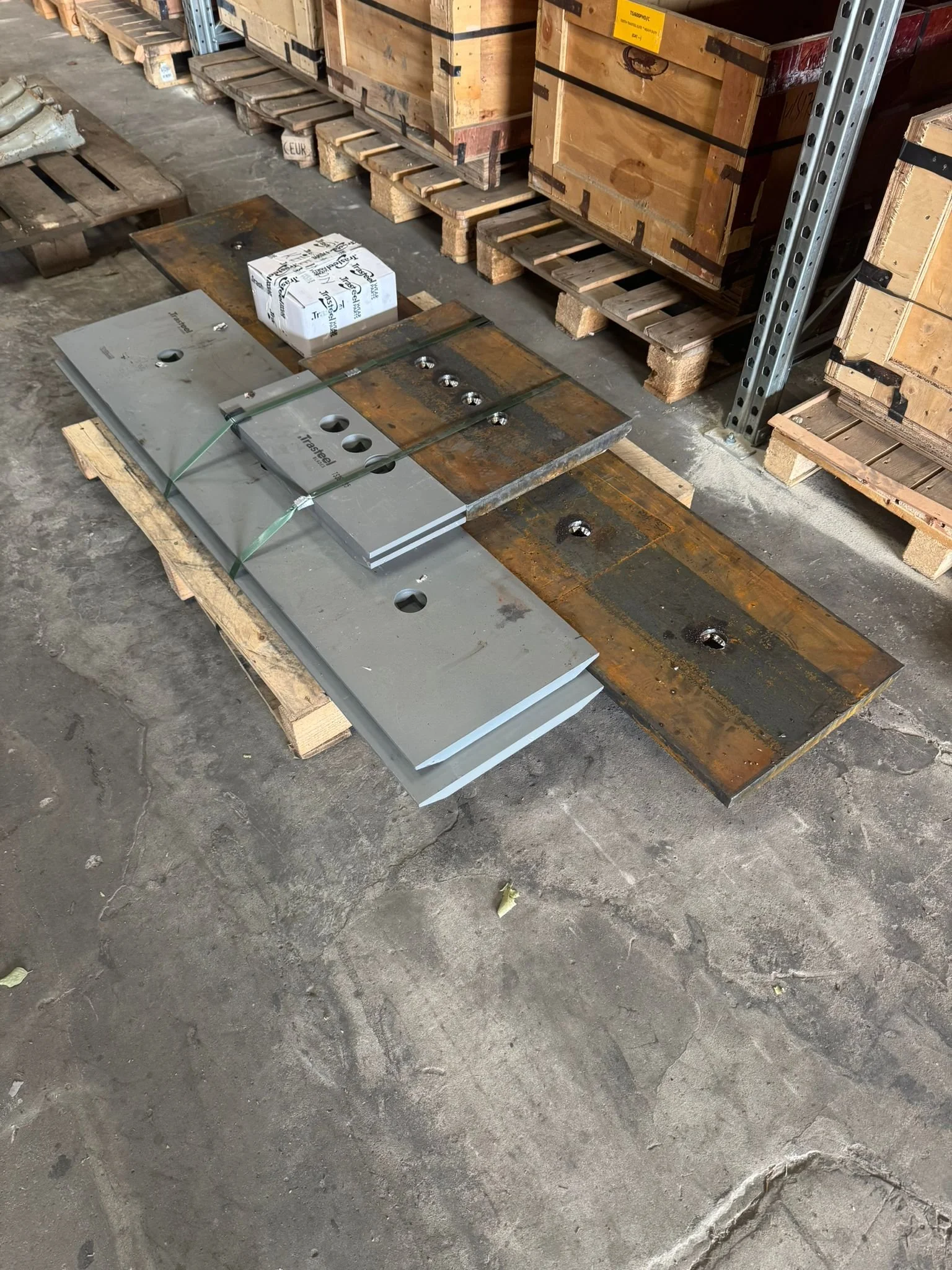 Steel plates and a small cardboard box on a wooden pallet in an industrial warehouse setting.