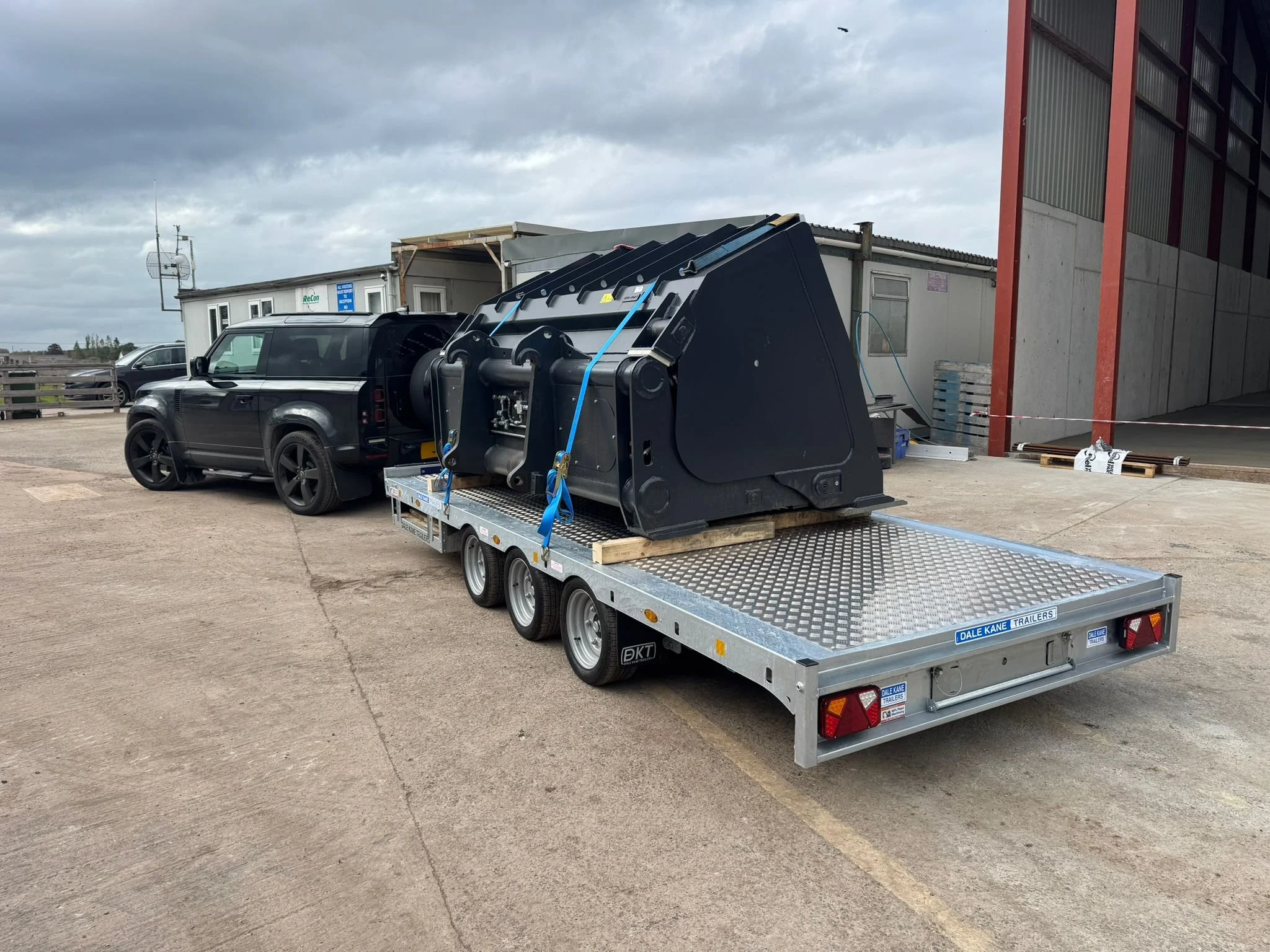 A black vehicle towing a flatbed trailer with a large black mechanical component secured on it, parked outside a building under cloudy skies.