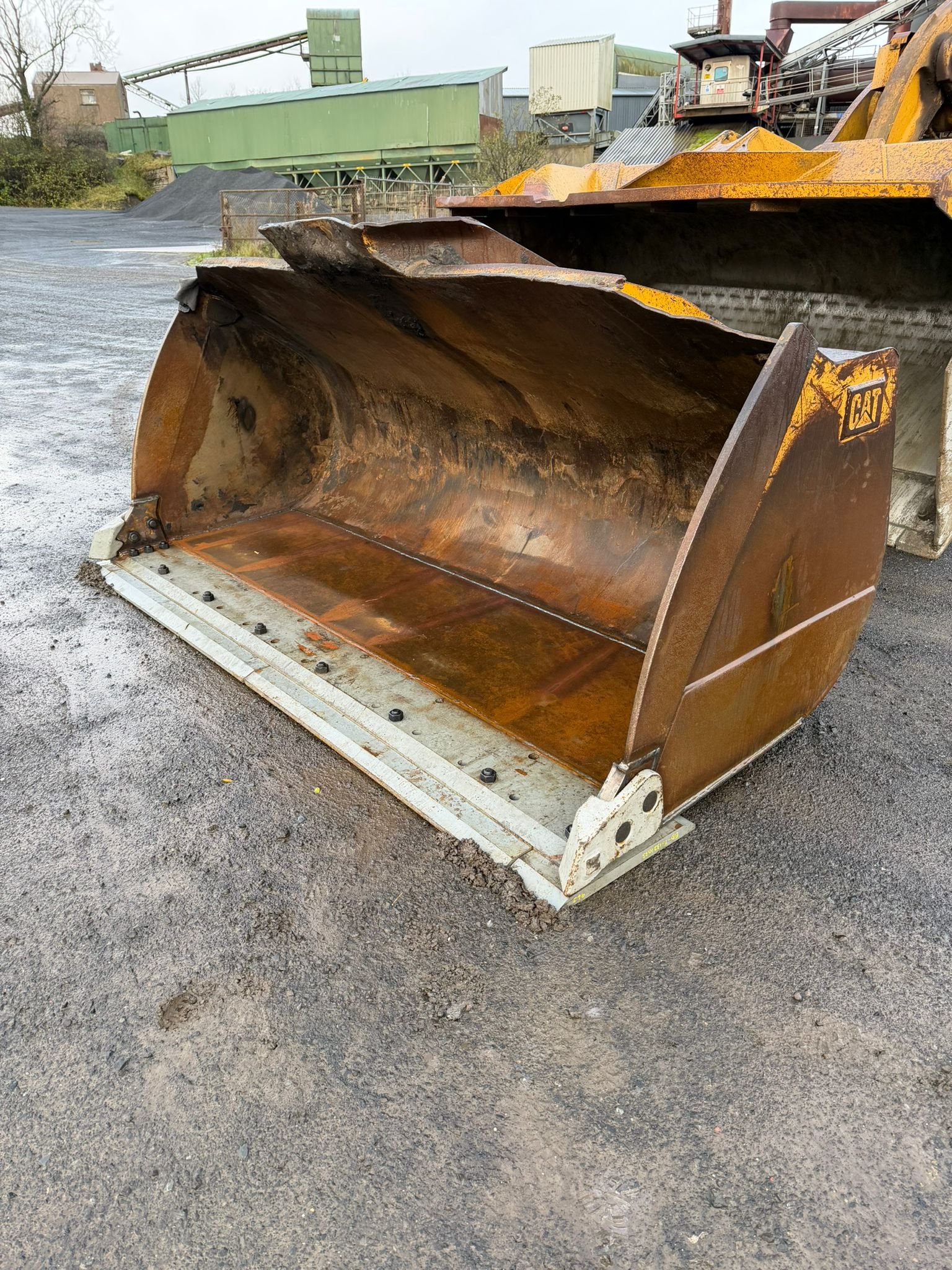 Rusty excavator bucket attachment on asphalt surface in an industrial area with factory buildings and equipment in the background.