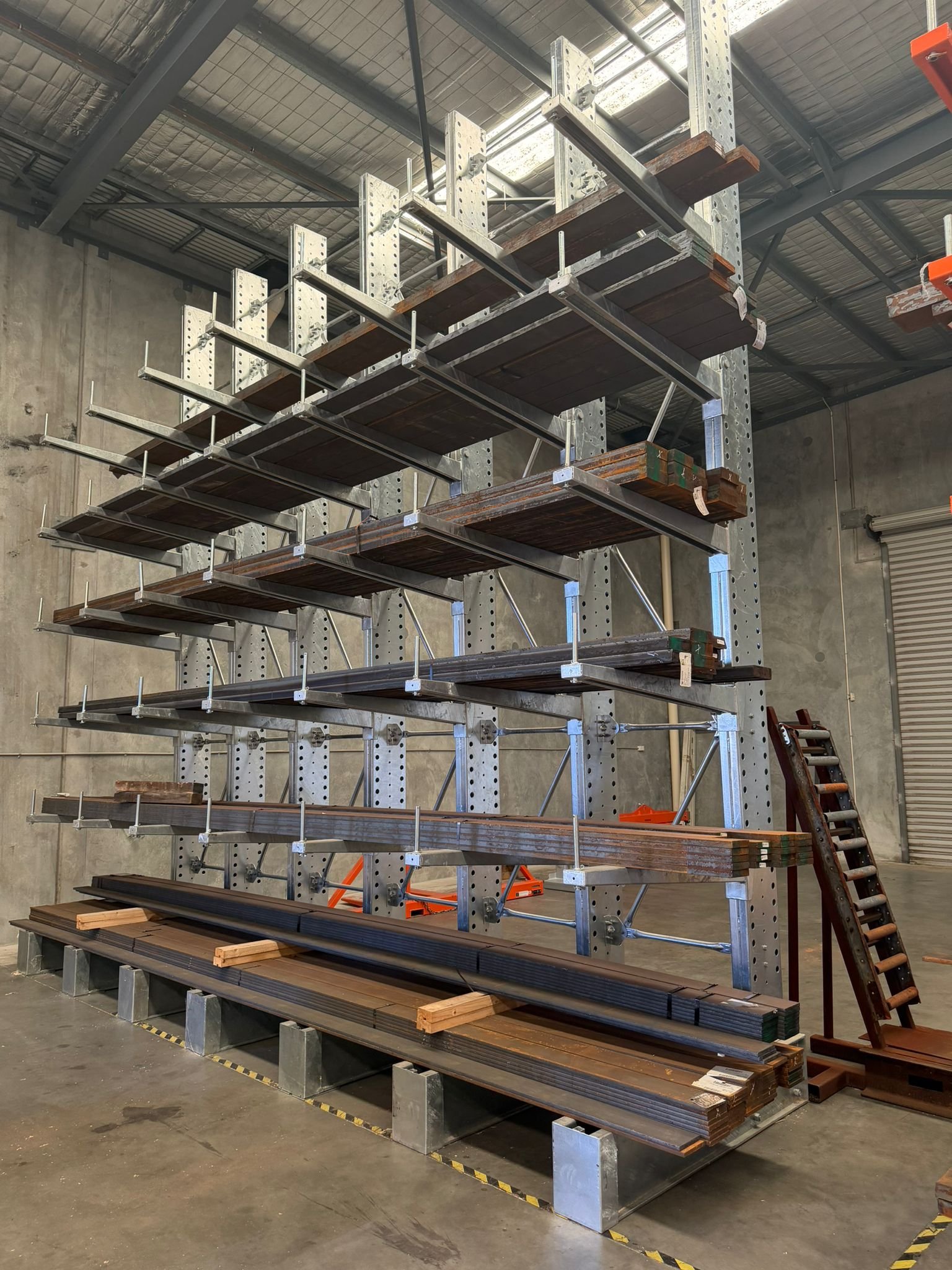 A metal storage rack with wooden planks and long metal bars stored on its multiple shelves inside a warehouse.