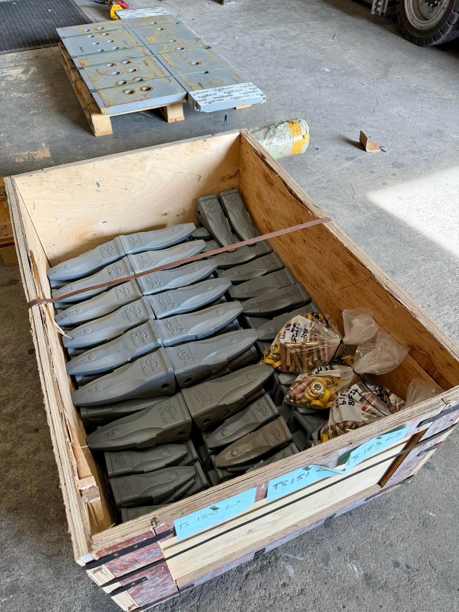 A wooden crate filled with gray electrical connectors and plastic bags of small brass fittings or fittings, on a concrete floor in a warehouse or workshop.