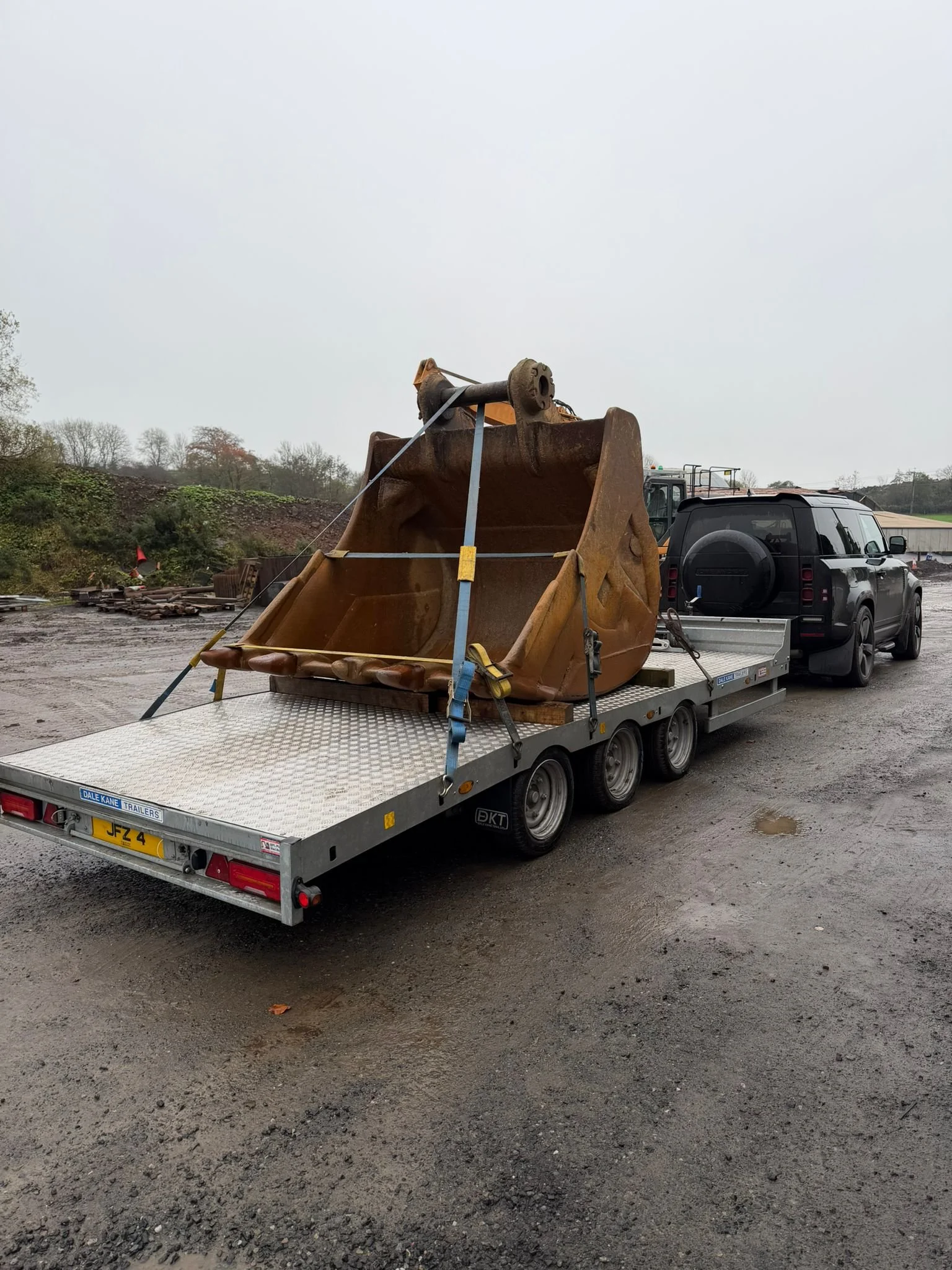 A large excavator bucket loaded on a flatbed trailer attached to a black SUV.