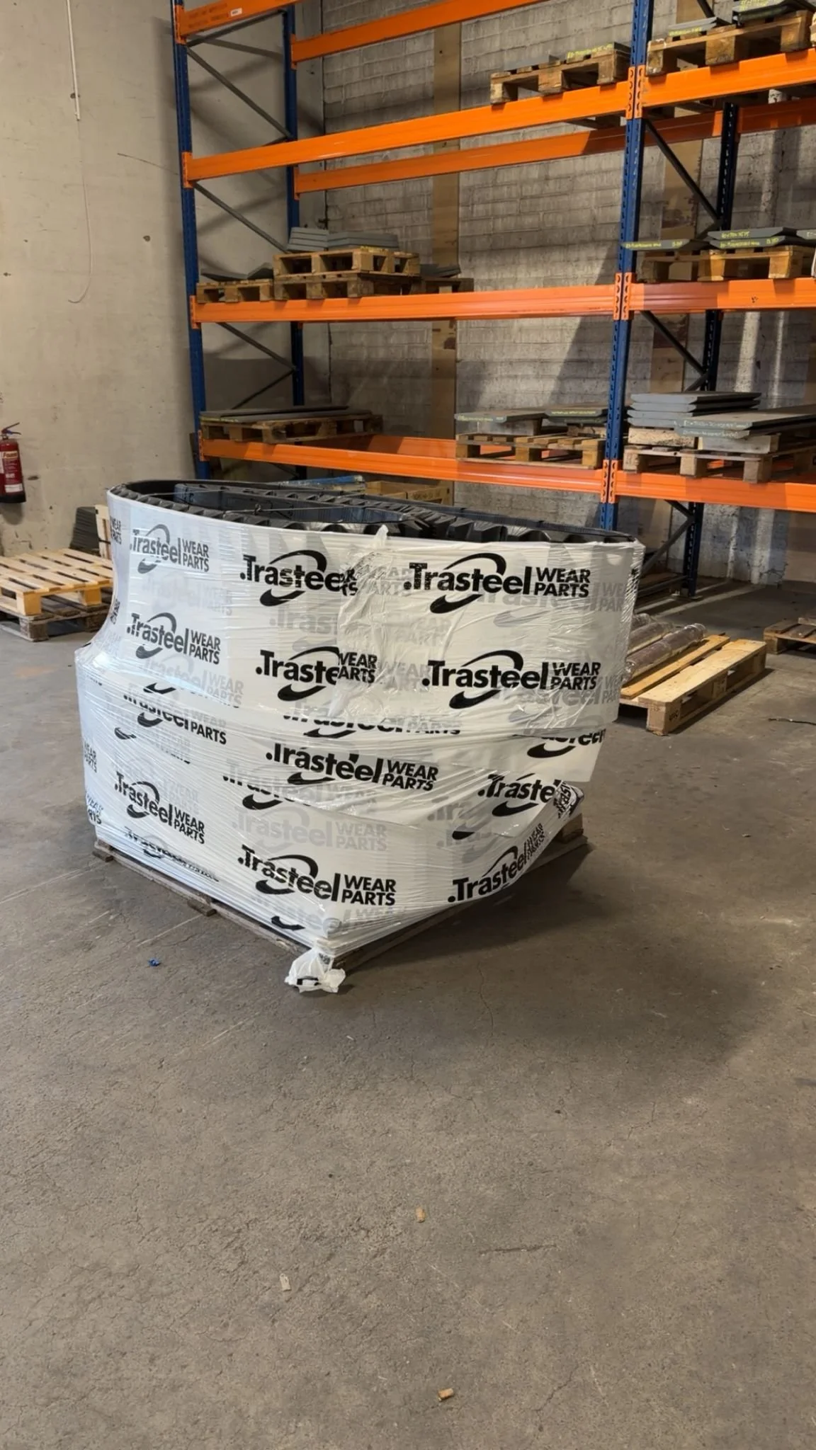 A pallet of wrapped Trasteel wear parts in a warehouse with orange and blue metal shelving units in the background.