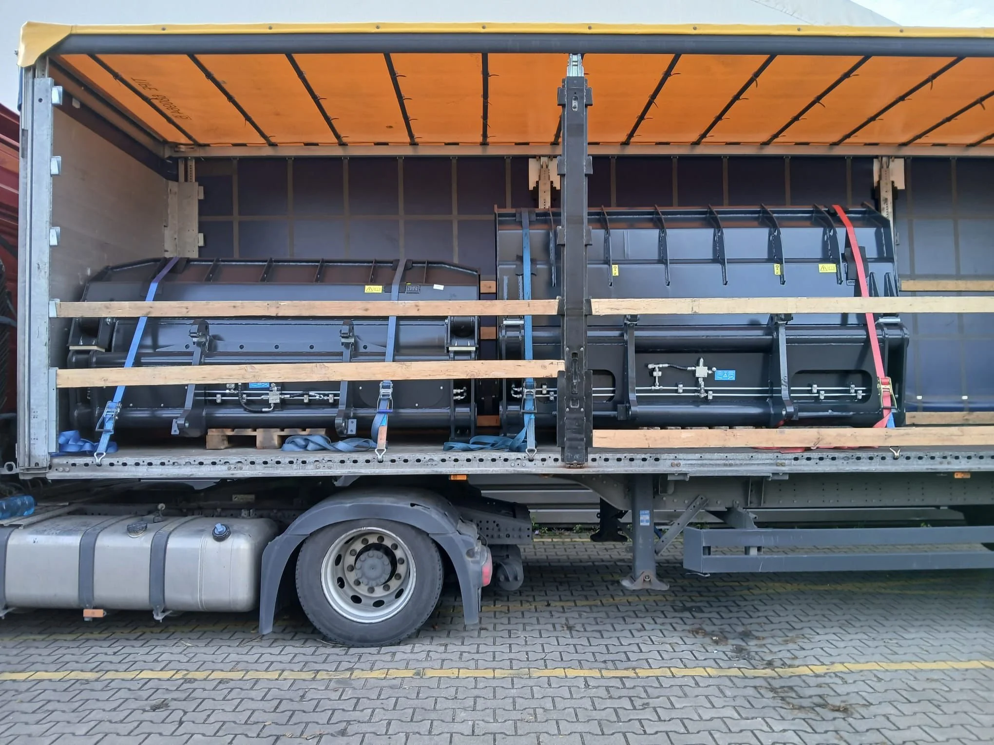 A truck with an open cargo area containing two large black cylindrical industrial tanks secured with straps and wooden beams.