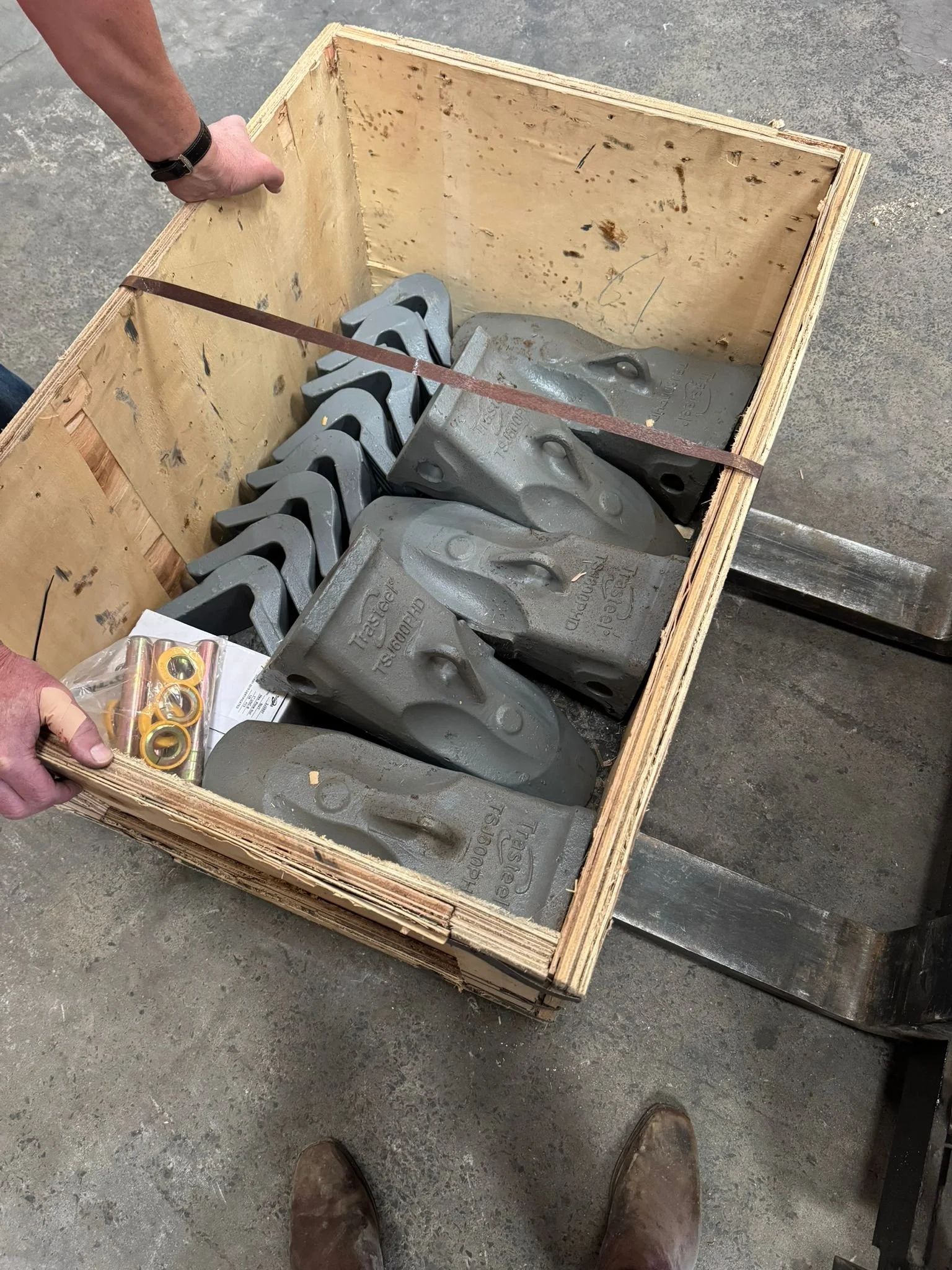 A wooden crate containing large steel excavator or bulldozer bucket teeth and a small bag of replacement bushings, on a concrete floor in a workshop or construction site.