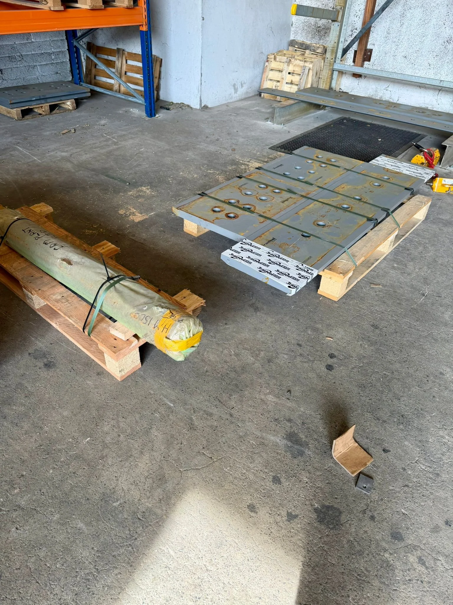 Construction materials on pallets, including a wrapped metal pipe and a metal ramp or platform, in a garage or warehouse setting.