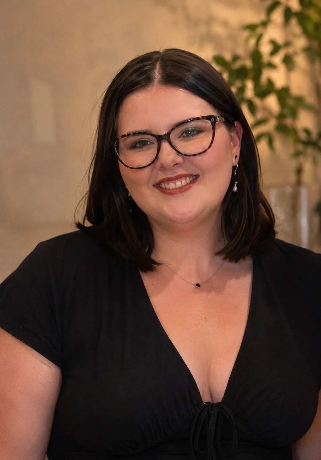 A woman with shoulder-length dark hair, glasses, and earrings, smiling indoors.