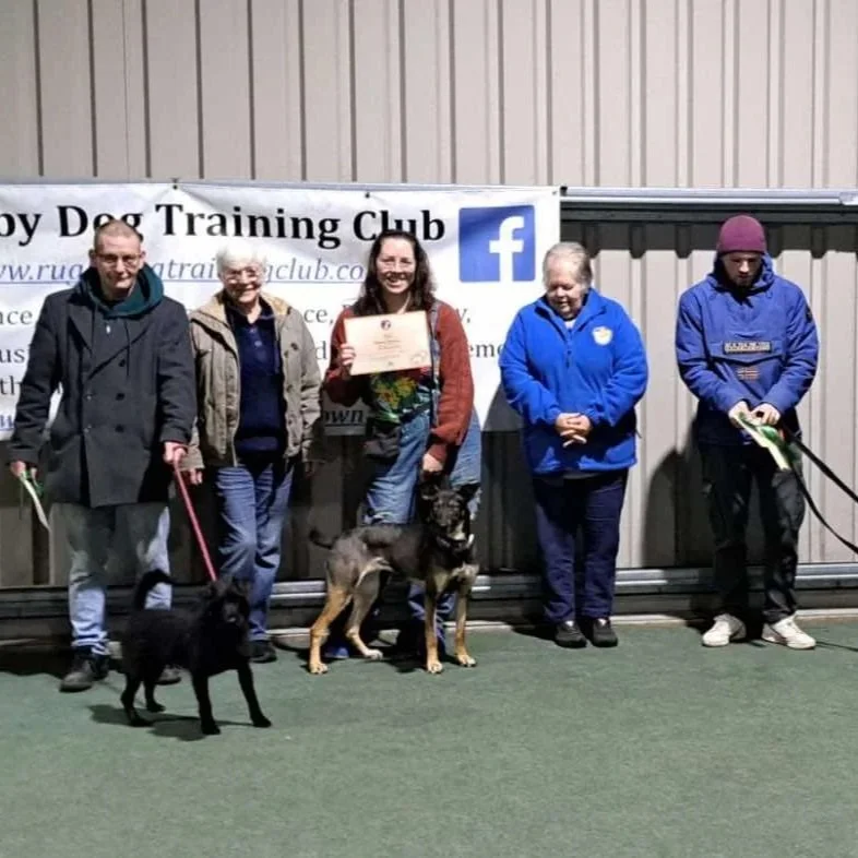 Group of seven people standing in front of a banner for Rugby dog training club, with five dogs on leads. Two of the people are holding a certificate and a rosette.