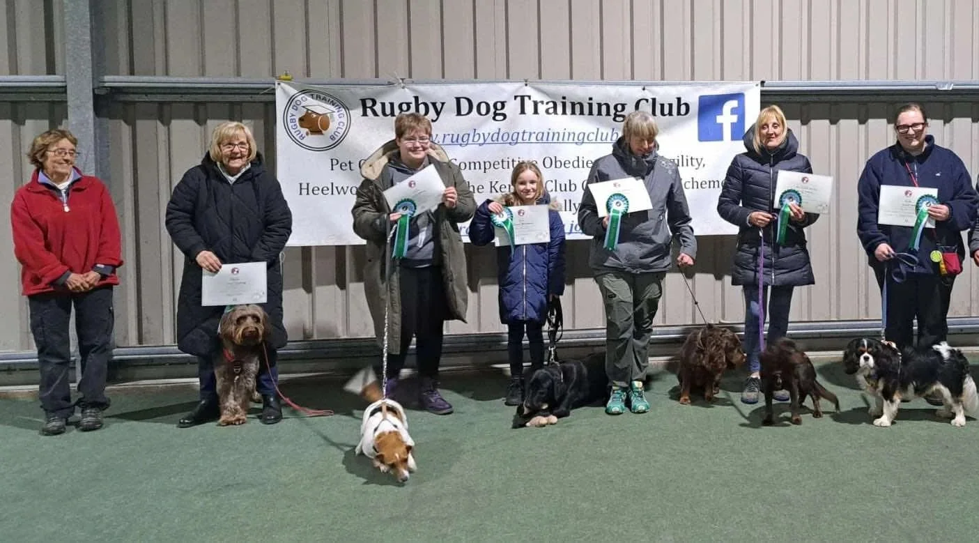 Group of people standing with dogs and holding certificates in front of a banner for Rugby Dog Training Club.