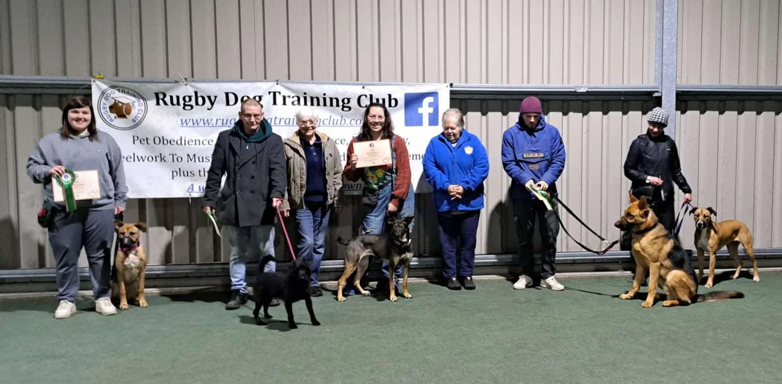 Group of people standing with their dogs in an indoor training facility, holding certificates and ribbons, with a banner reading 'Rugby Dog Training Club' behind them.