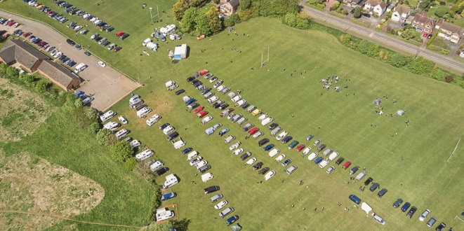 Aerial view of Rugby DTC Obedience show, including a large field filled with cars, next to a grassy field with people and some structures nearby.