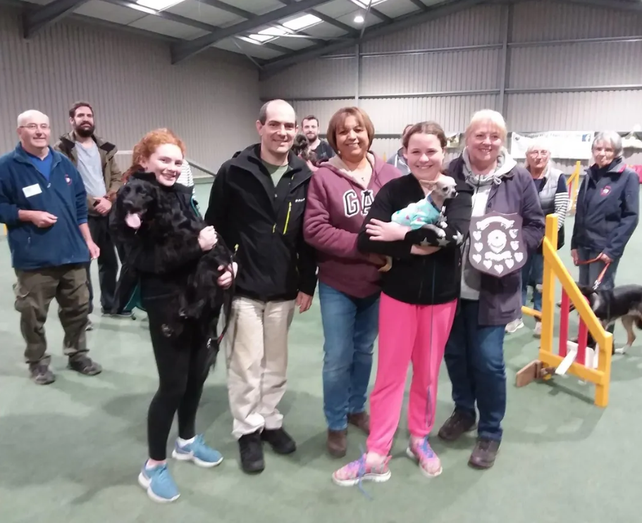Group of people inside a large indoor facility, some holding dogs, with agility equipment in the background, celebrating.