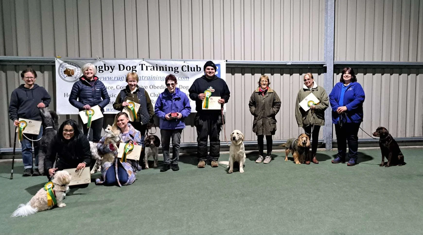 Group of people and their dogs at a Rally Dog Training Club event, holding certificates and ribbons, indoors on a green carpet with a banner in the background.