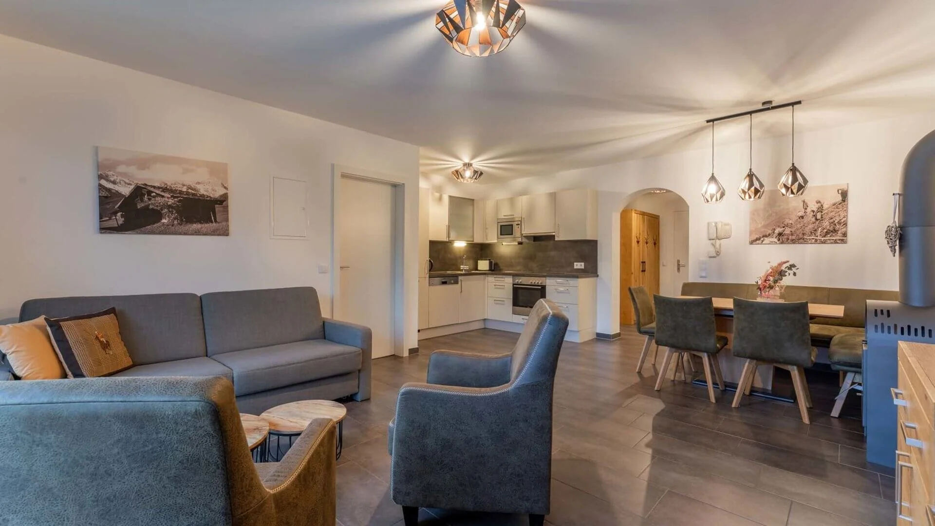 Offener Wohn- und Essbereich eines modernen Apartments von eder VIEW in Maria Alm mit grauem Sofa, Sesseln, Esstisch mit Stühlen, moderner Küche im Hintergrund, Deckenleuchten und Wandbilder im skandinavischen Stil.