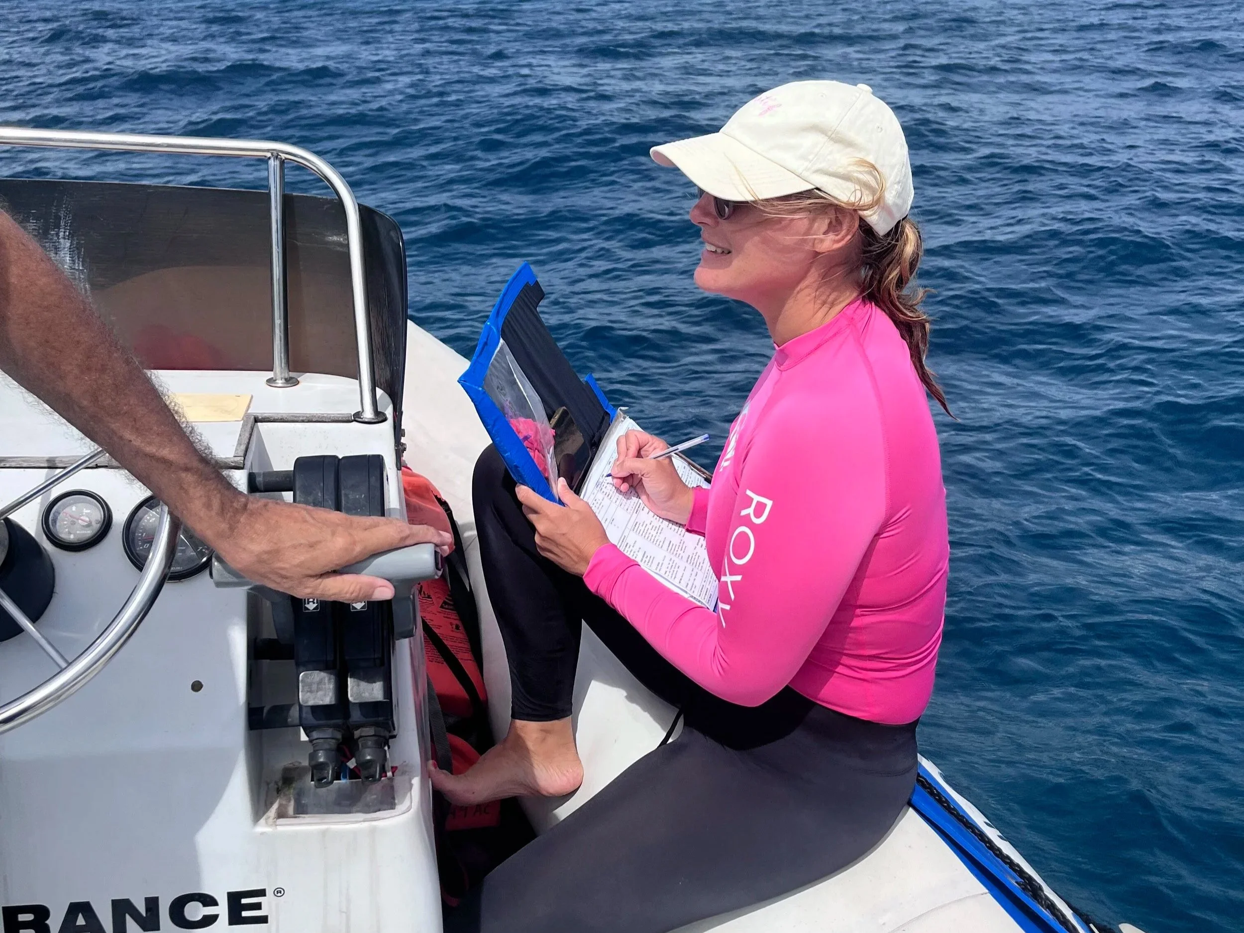 Laure souriante portant une casquette blanche et un haut de plongé rose, assise dans un bateau en pleine mer au Mozambique, en train de prendre des notes.