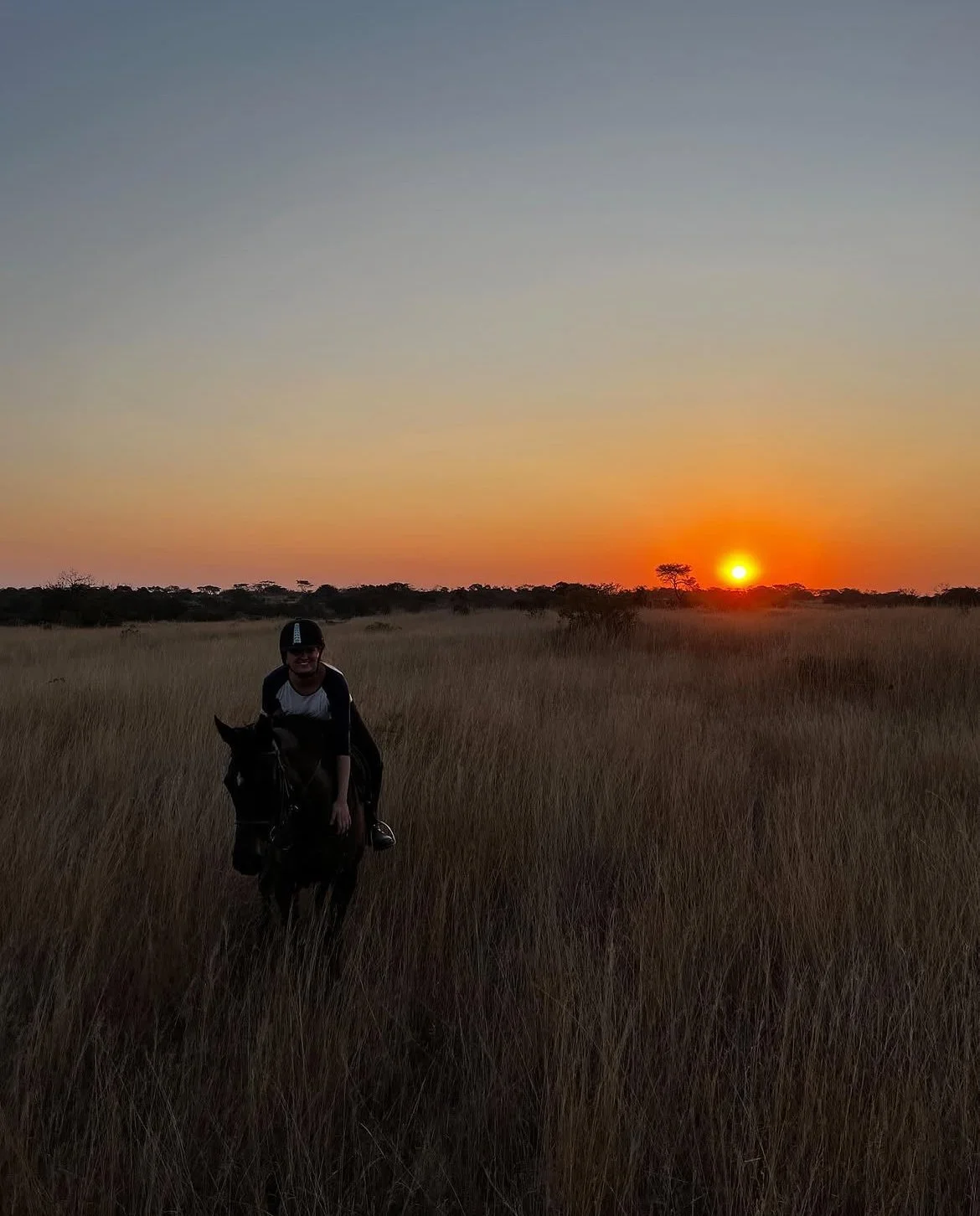 Une personne en chemise noire et blanche chevauche un cheval dans un champ d'herbe haute au coucher du soleil, avec un ciel dégagé en arrière-plan.