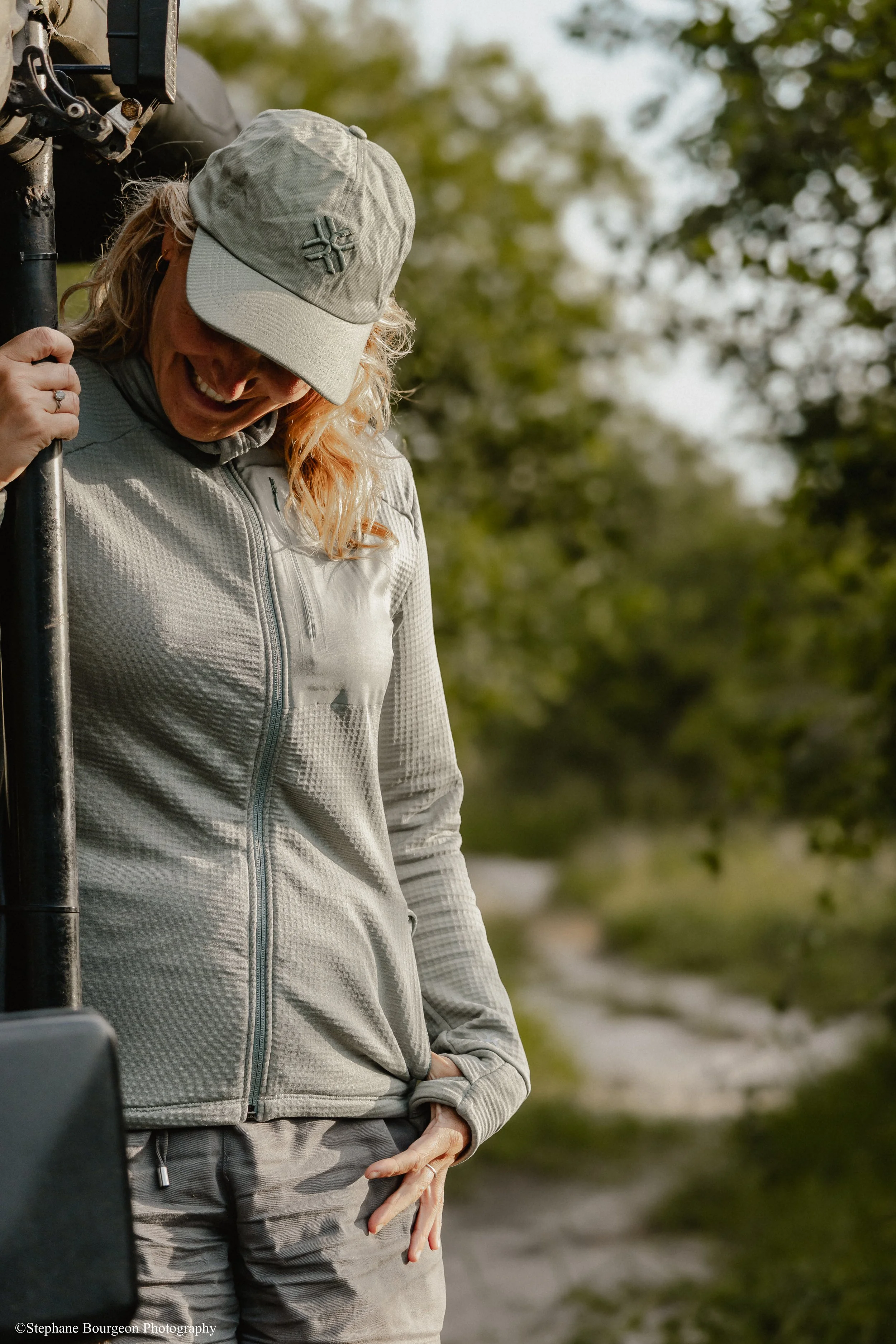 Laure souriante portant une casquette beige et une veste grise, debout à l'extérieur parmi des arbres, avec une main dans la poche, apparemment sous une lumière naturelle douce.