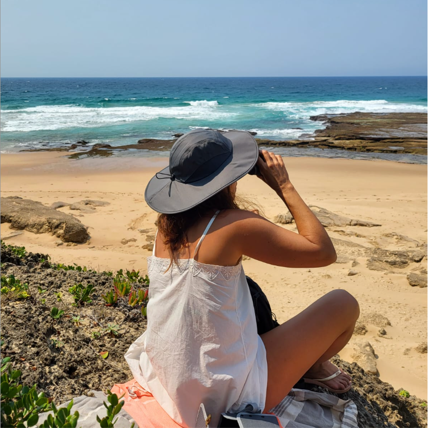 Une femme assise sur une roche face à la mer, portant un chapeau gris et un vêtement blanc, regardant l'océan.