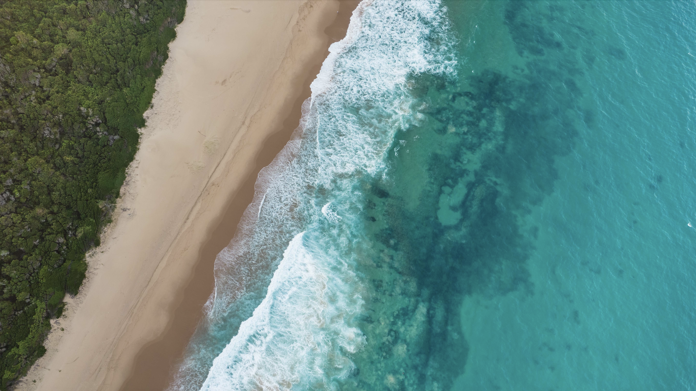 Vue aérienne d'une plage avec du sable beige au Mozambique, des vagues turquoise et une zone de végétation verte dense à gauche.