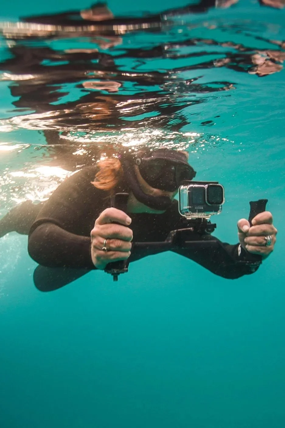 Une personne en plongée sous-marine, portant un masque et une combinaison noire, tient une caméra ou une action cam sous l'eau.