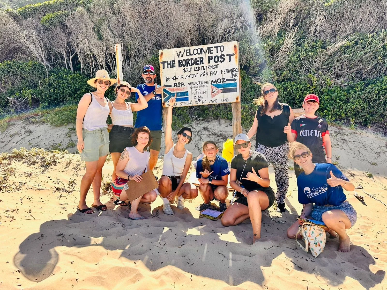 Groupe de personnes posant sur la plage près d'un panneau indiquant l'entrée du poste frontière avec des touristes portant des chapeaux, des lunettes de soleil, certains tenant des documents, et tous souriant ou faisant des gestes de bonne humeur.