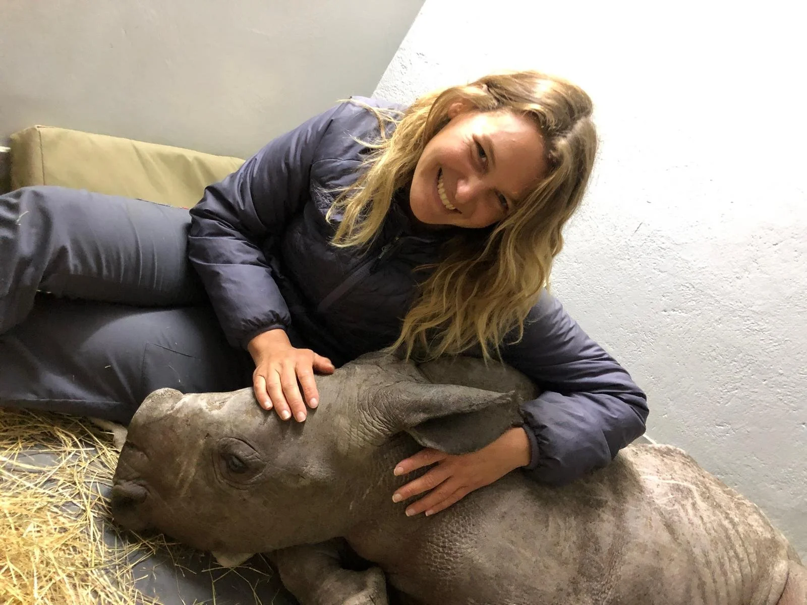 Une femme souriante couche horizontalement, caressant un bébé rhinocéros, dans un environnement intérieur avec un mur blanc et de la paille au sol.