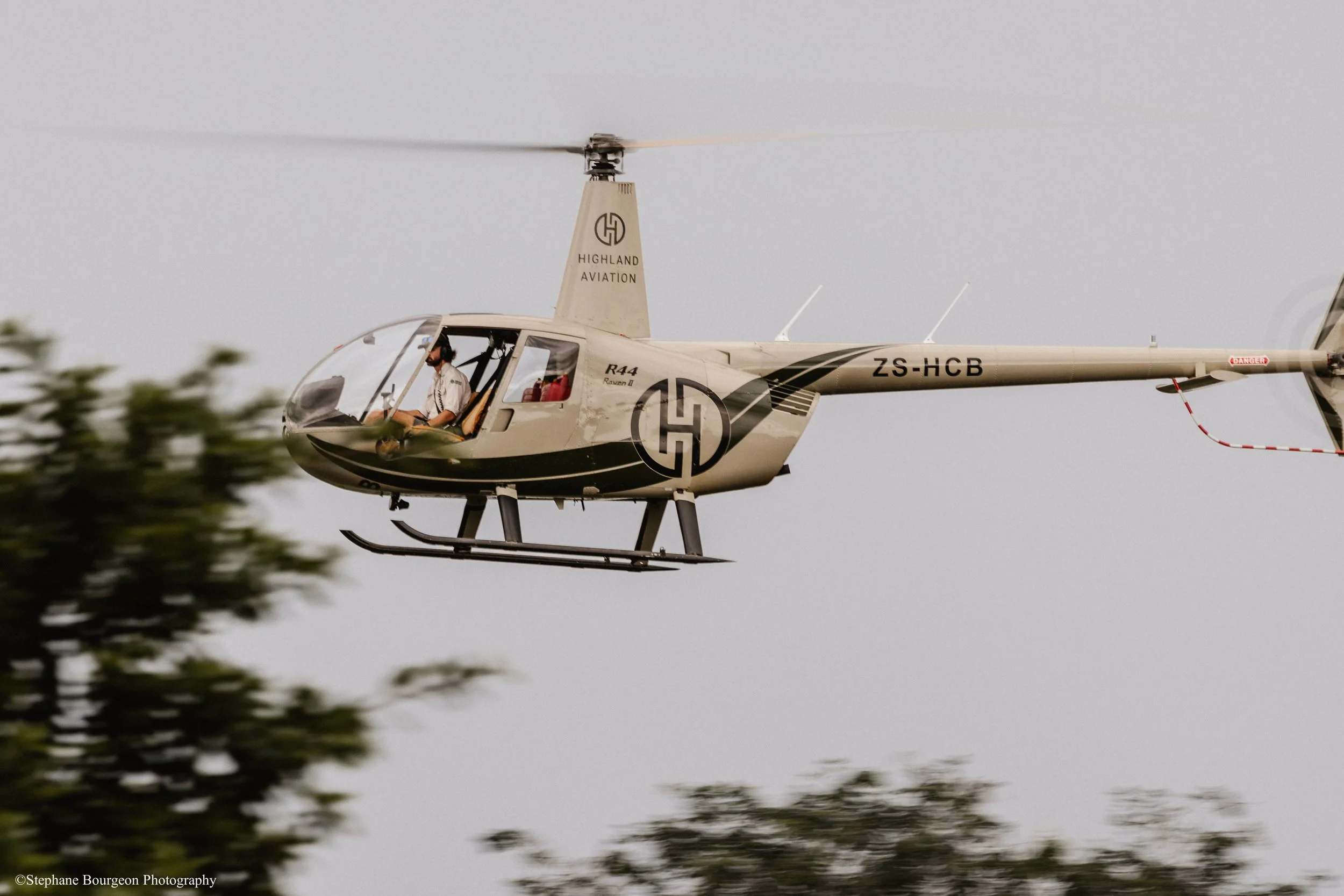 Un hélicoptère de la marque Highland Aviation en vol dans le ciel, avec un pilote visible dans la cabine. Des arbres partiellement flous apparaissent en bas de l'image.