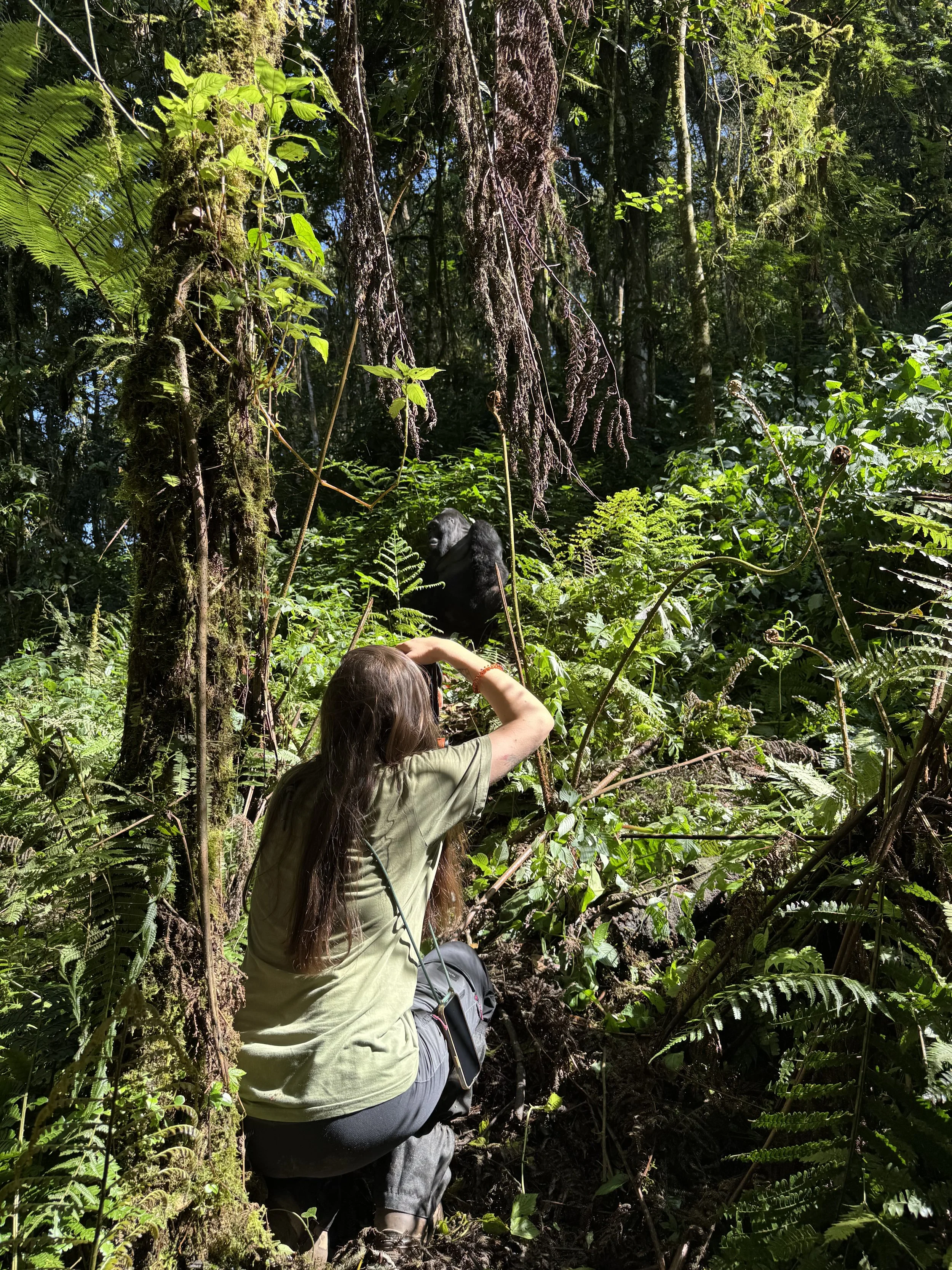 Une femme avec des cheveux longs bruns, portant un t-shirt vert, prend une photo d'un gorille dans la forêt dense.