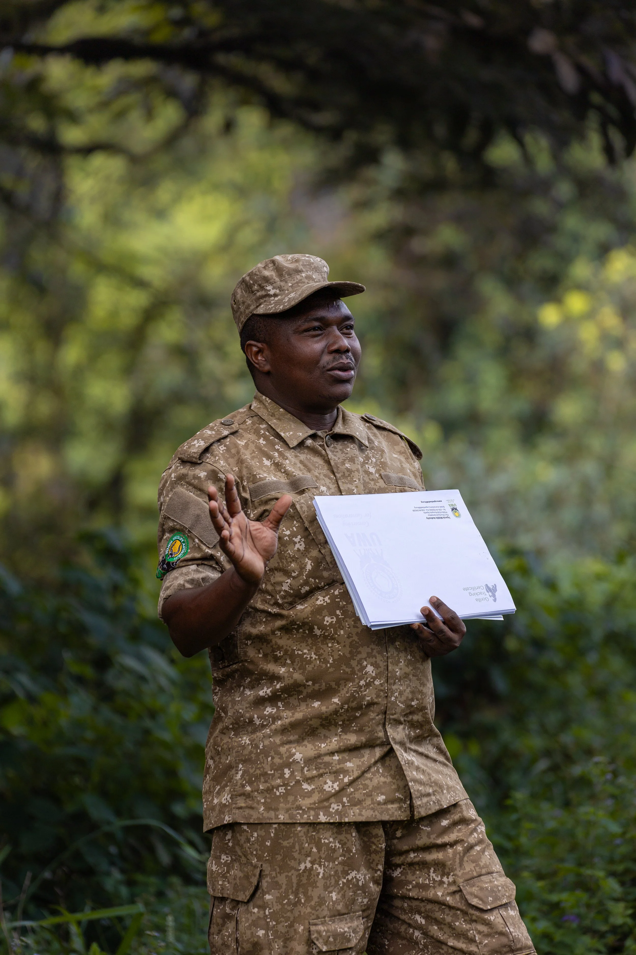 Un homme en uniforme militaire en train de parler, tenant un document, dans un environnement naturel avec des arbres en arrière-plan.