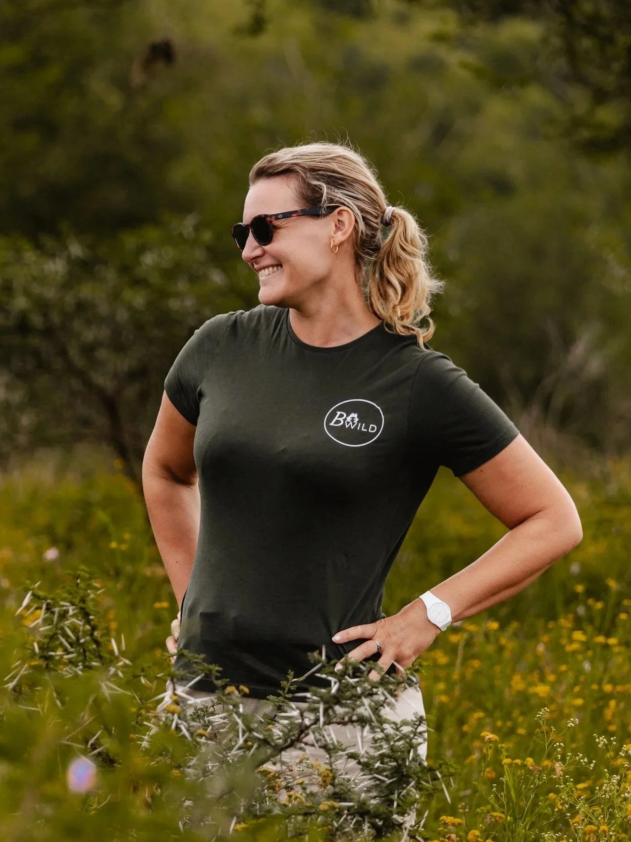 Laura souriante portant des lunettes de soleil, un t-shirt vert avec le logo blanc de l'association Bwild, dans un environnement naturel avec des arbres et des fleurs en Afrique du Sud.
