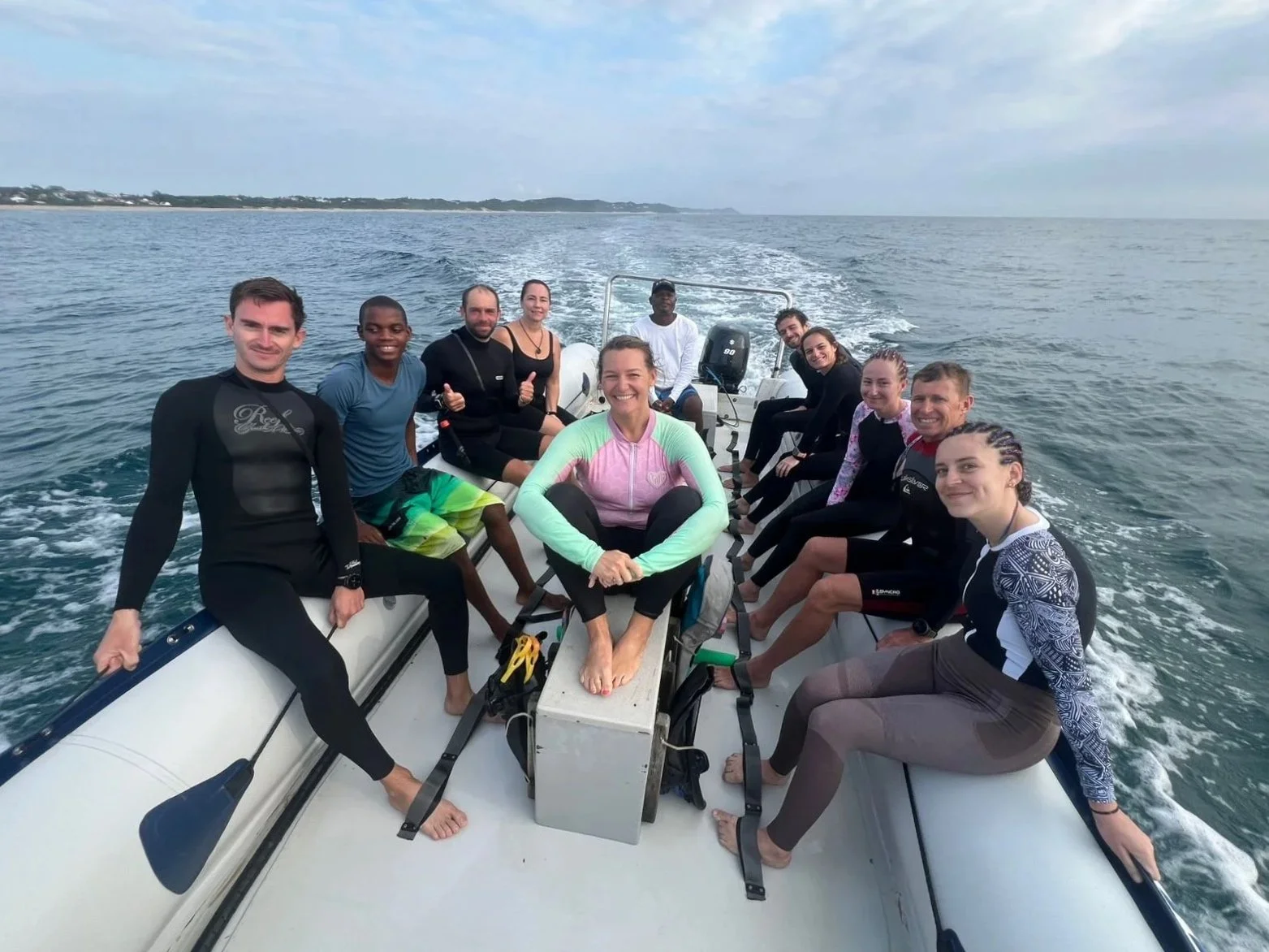 Groupe de personnes à bord d'un bateau en mer, quelques-uns en maillot de bain, certains souriants, paysage marin avec horizon et côte en arrière-plan.